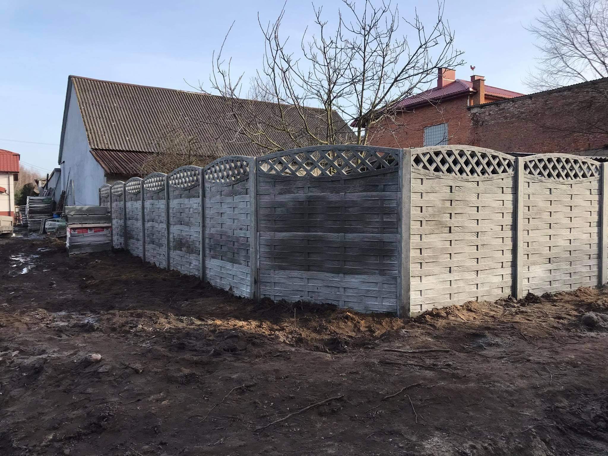 Nowo postawione, betonowe ogrodzenie panelowe o wzorze imitującym plecionkę, na tle zaoranego terenu i budynków z różnymi pokryciami dachowymi.