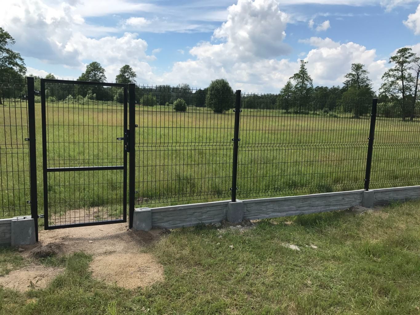 Ciemna, metalowa siatka ogrodzeniowa z furtką na betonowej podmurówce, widok na łąkę i las w tle pod błękitnym niebem z chmurami.