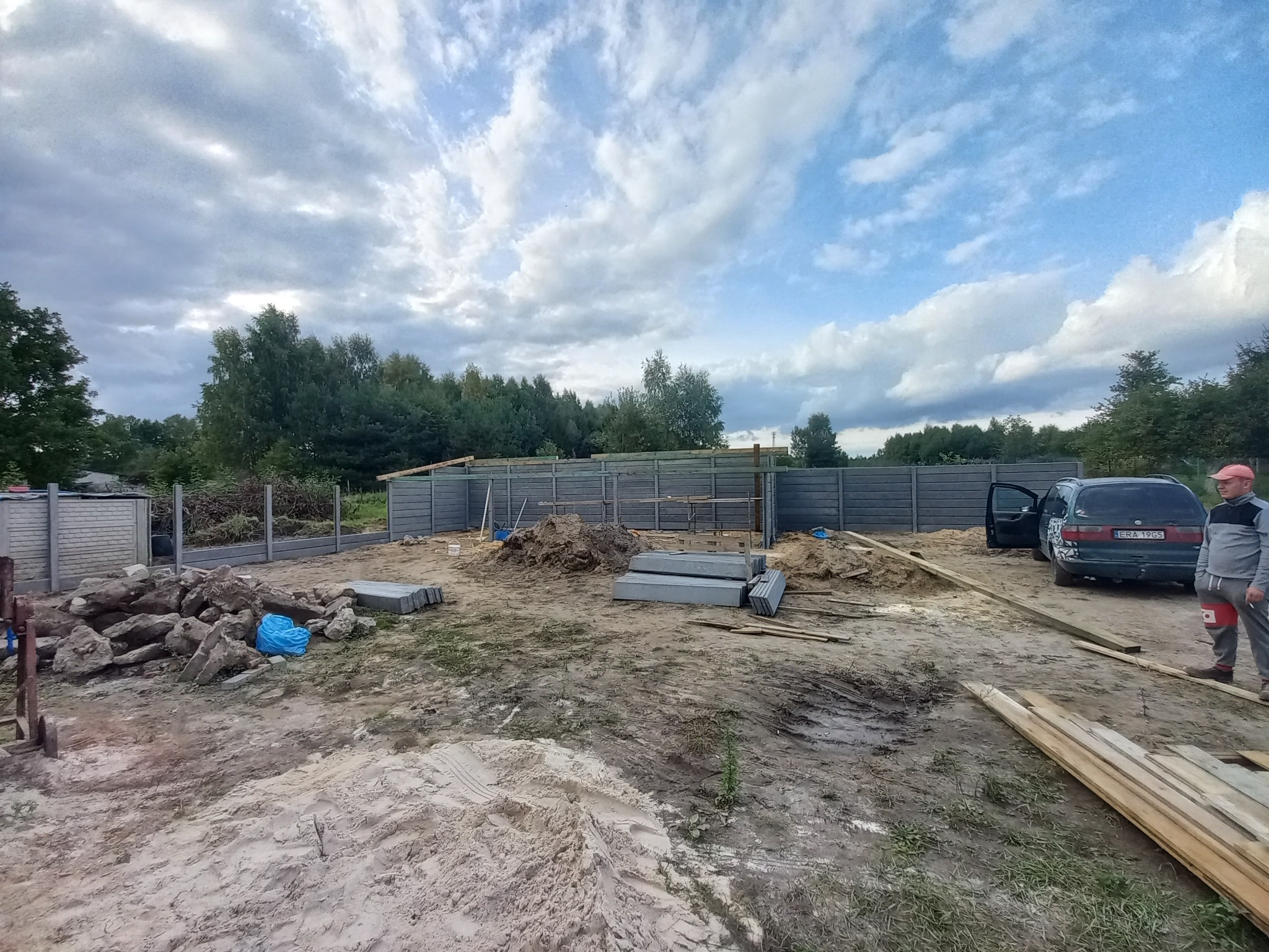 W trakcie budowy betonowego ogrodzenia panelowego z widocznymi elementami fundamentu, stertą ziemi, deskami i samochodem osobowym z otwartymi drzwiami w tle, pochmurne niebo.