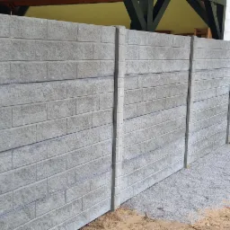 Szary, betonowy mur imitujący cegłę, zakończony świeżo wysypanym drobnym żwirem, z drewnianym płotkiem i stertą drewna w tle.