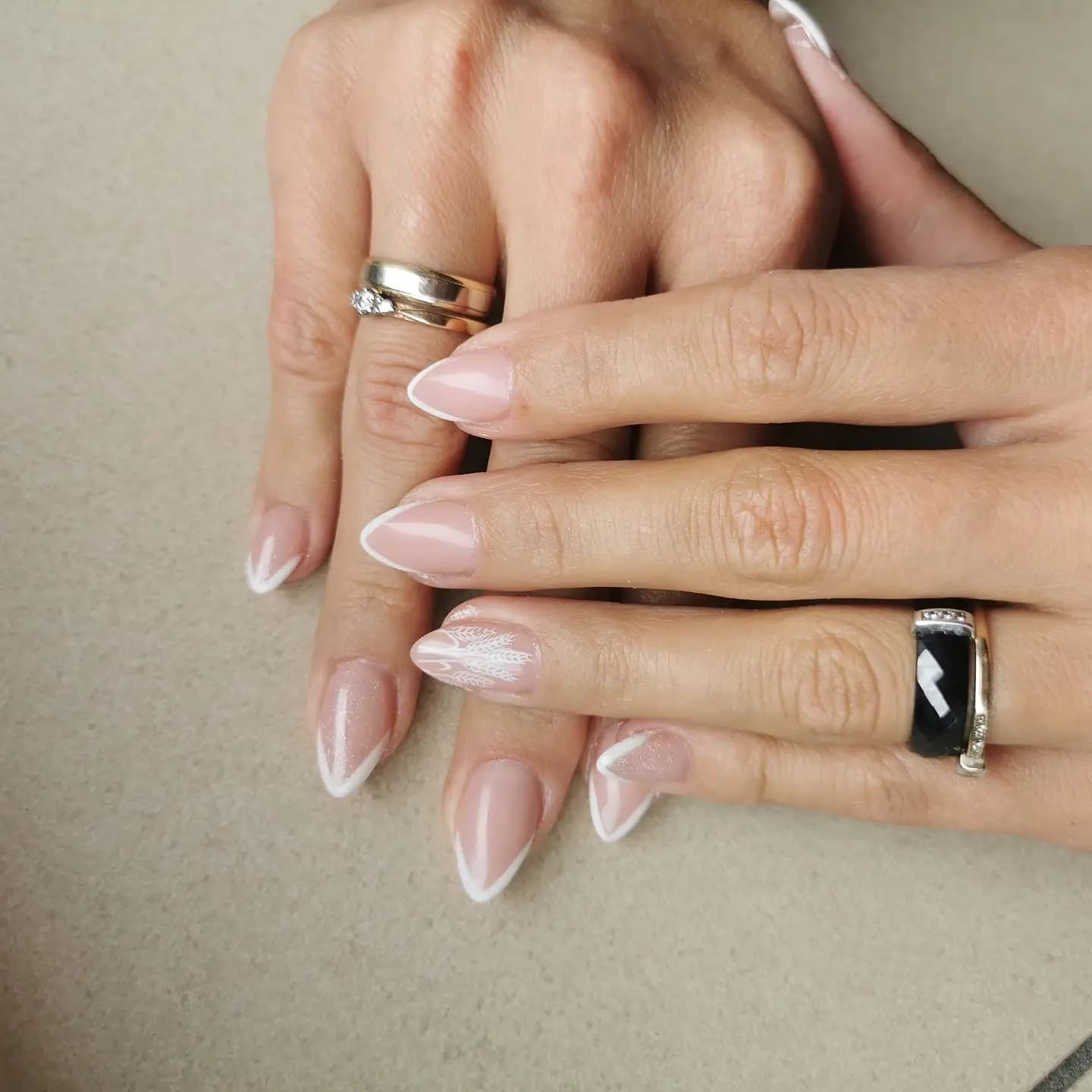 Dłonie z jasnym manicure francuskim i delikatnym białym zdobieniem w kształcie piórka na jednym paznokciu, pierścionki na palcach.