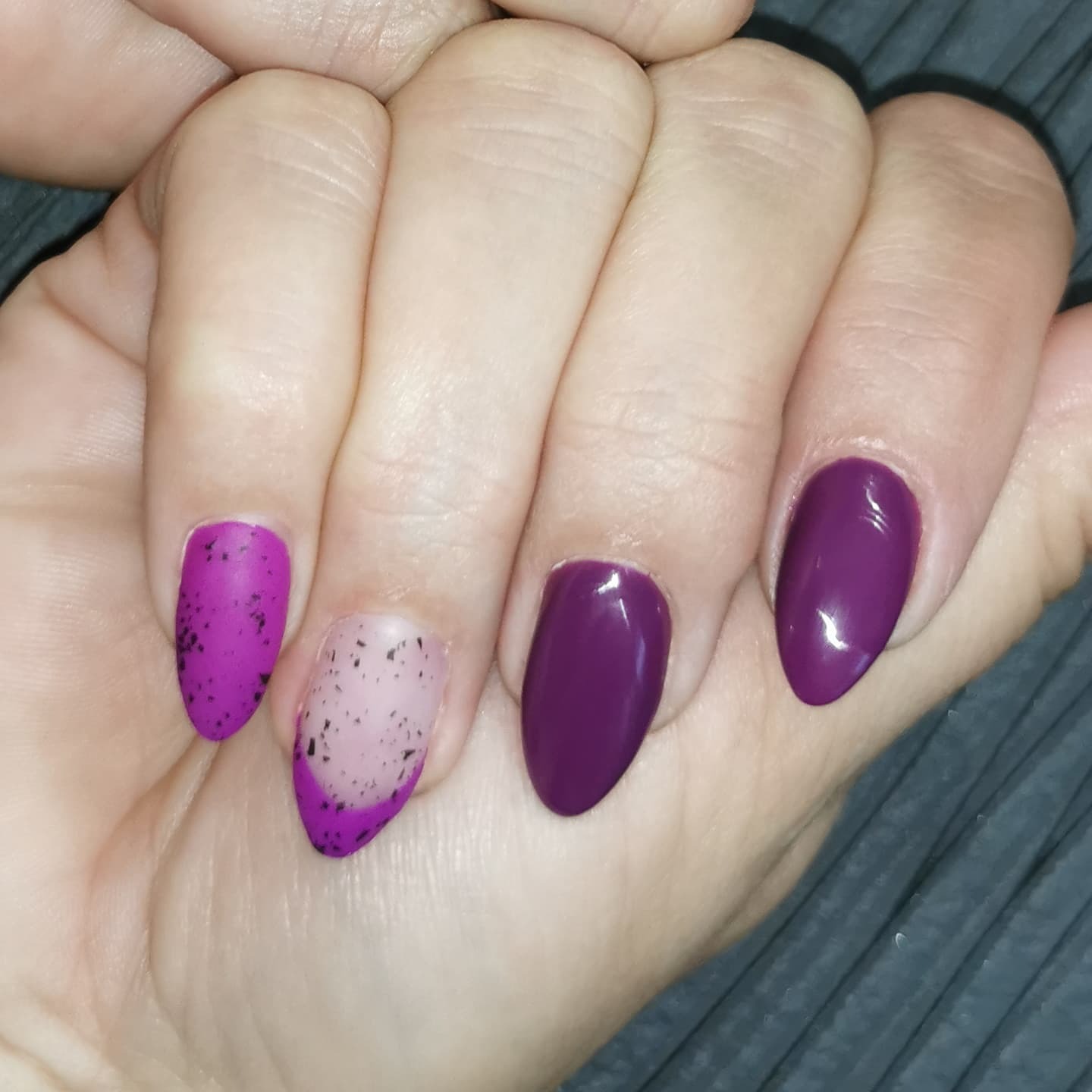 Dłoń z pomalowanymi paznokciami: dwa w odcieniach fioletu z czarnymi plamkami, dwa jednolite w kolorze śliwki, jeden z naturalną bazą i fioletowym półksiężycem u nasady.