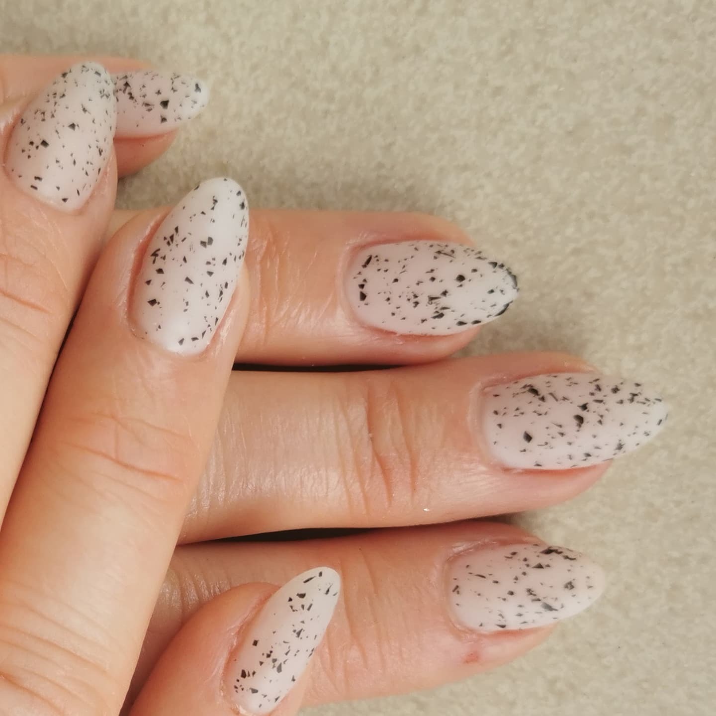 Dłoń z owalnymi paznokciami pomalowanymi na jasny kolor z czarnymi, nieregularnymi plamkami, imitującymi przepiórcze jajko.