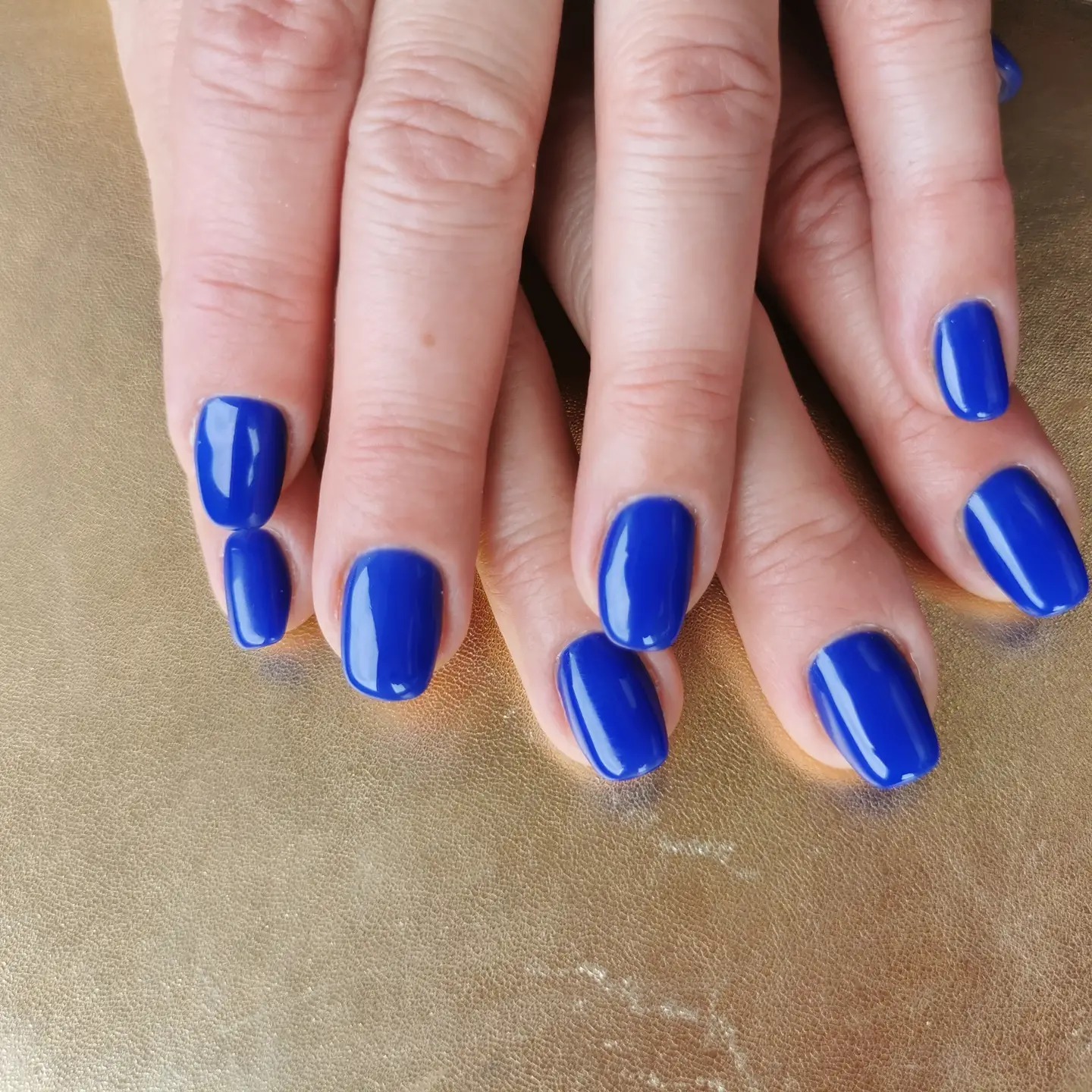 Dłonie z krótkimi, kwadratowymi paznokciami pomalowanymi na intensywny, błyszczący kobaltowy kolor, ułożone na złotej, teksturowanej powierzchni.