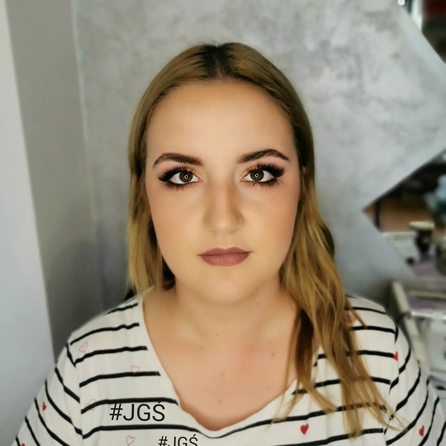 Portret kobiety z profesjonalnym makijażem oczu i ust w odcieniach brązu, ubranej w bluzkę w paski z drobnymi czerwonymi serduszkami, na rozmytym tle salonu.