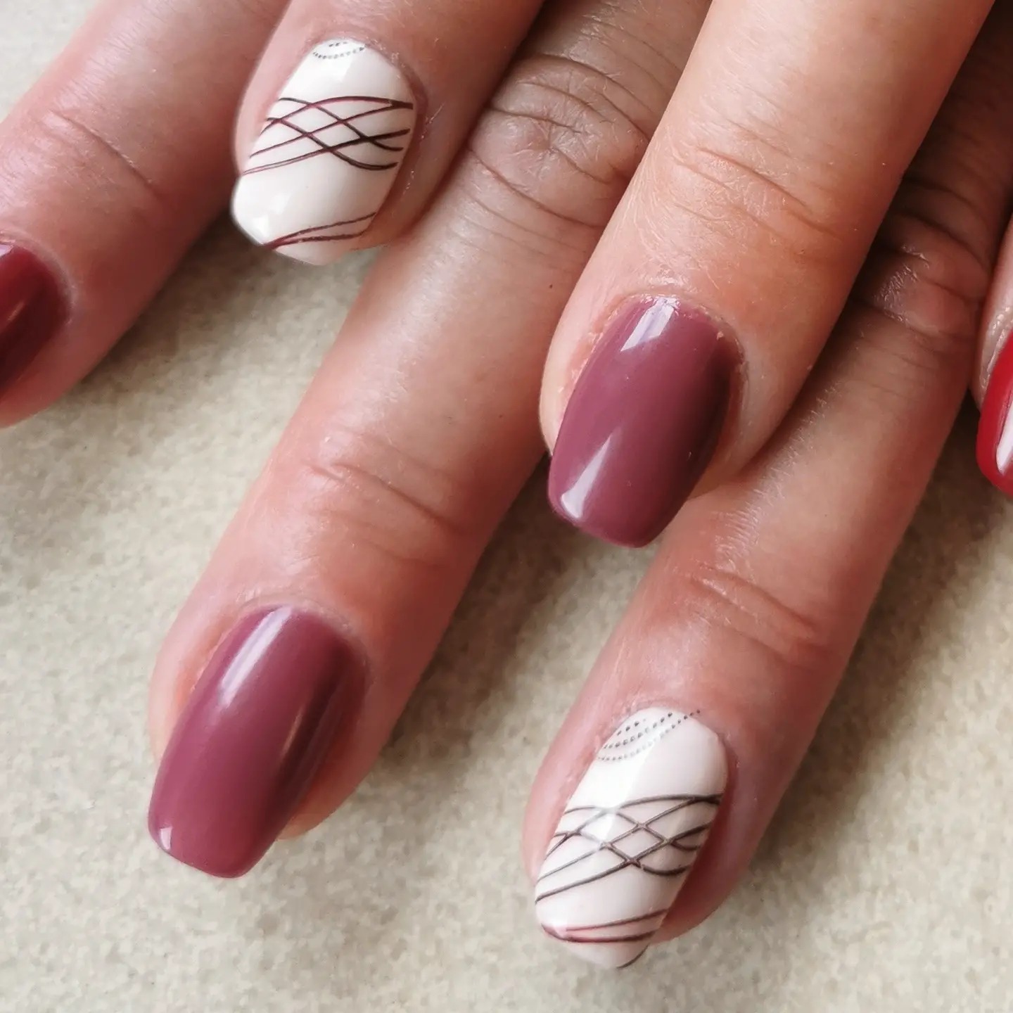 Dłoń z manicure hybrydowym: dwa paznokcie w kolorze burgundu, dwa białe z brązowymi liniami tworzącymi wzór siatki i drobnymi srebrnymi kropkami przy skórkach, jeden czerwony.