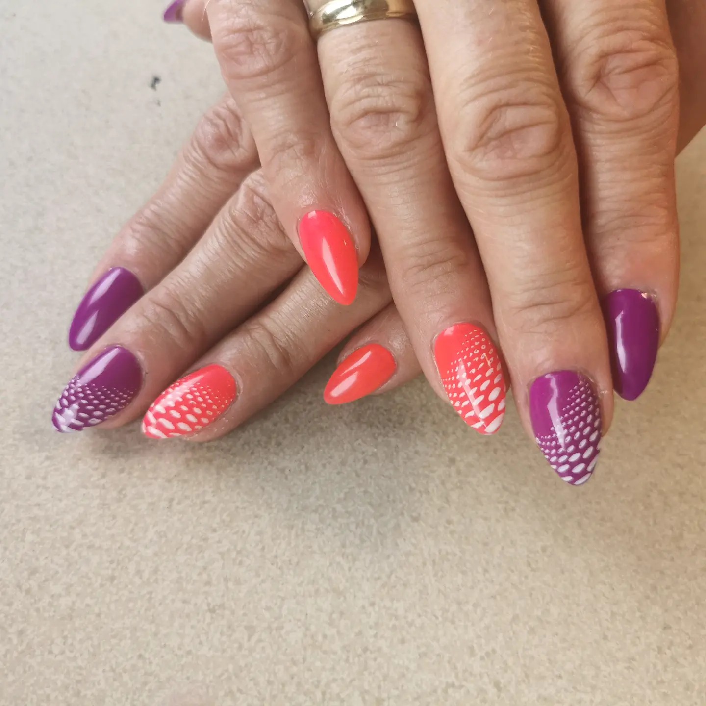 Dłonie z pomalowanymi paznokciami w kolorach fuksji i koralu, z białym wzorem kropek na niektórych paznokciach, na neutralnym tle.