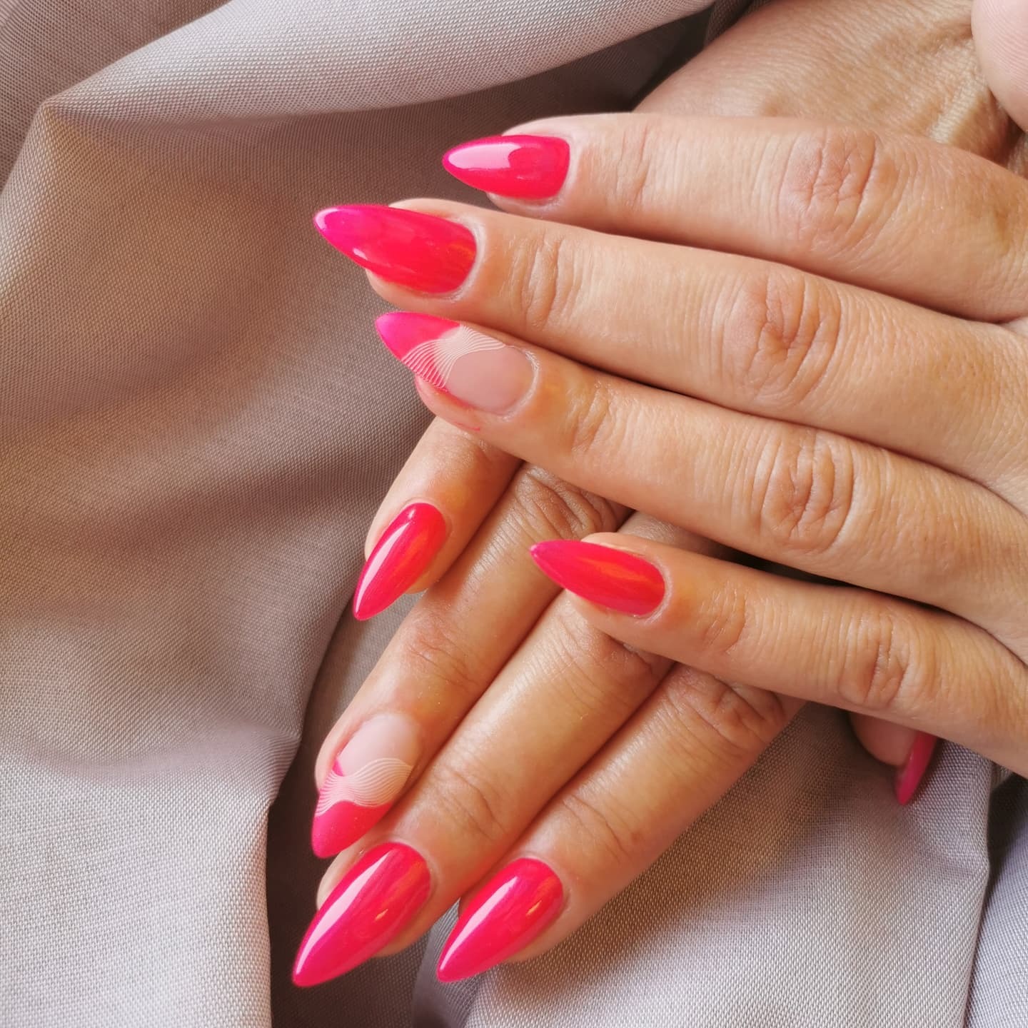 Dłonie z jaskrawo różowym manicure w kształcie migdała, jeden paznokieć z motywem fal na jasnym tle.