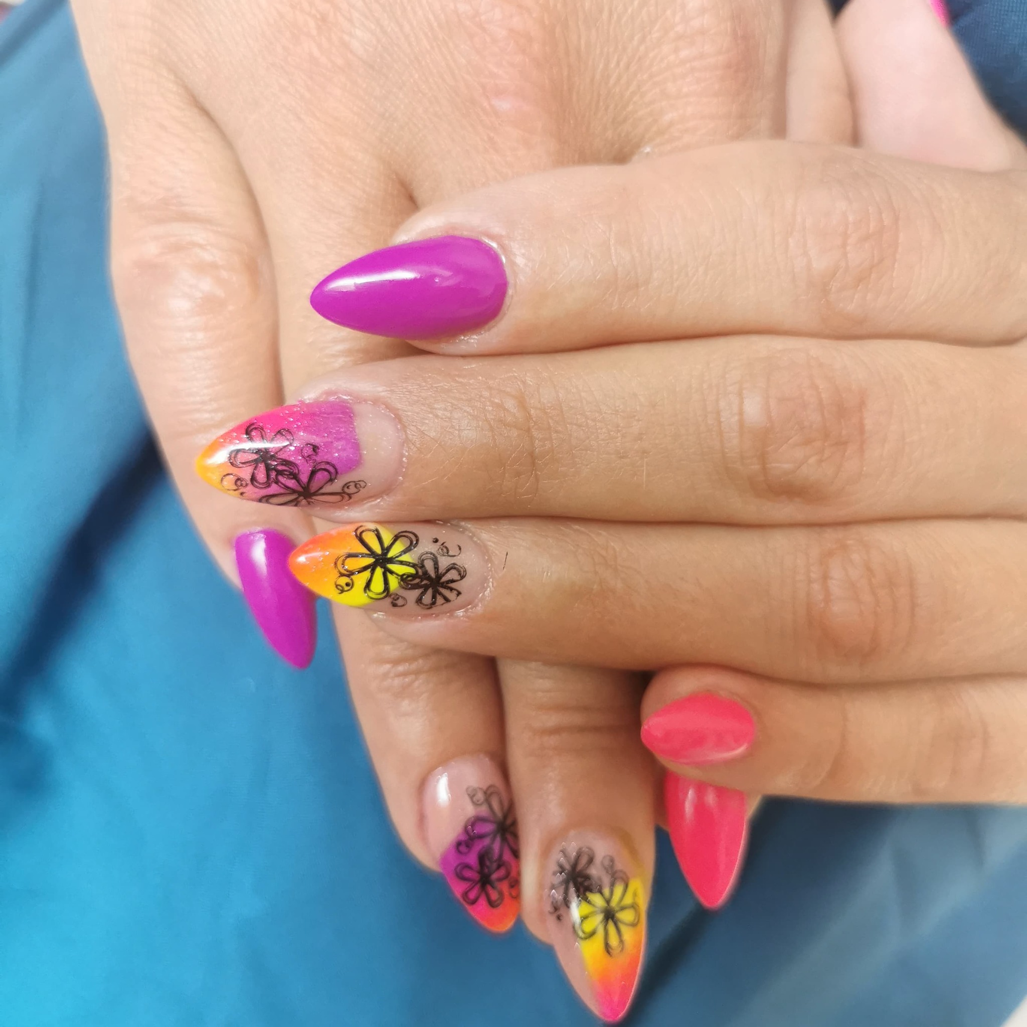 Dłonie z pomalowanymi paznokciami w odcieniach różu, pomarańczy i żółci, ozdobione czarnymi wzorami kwiatowymi na przezroczystej bazie, ułożone na niebieskim tle.