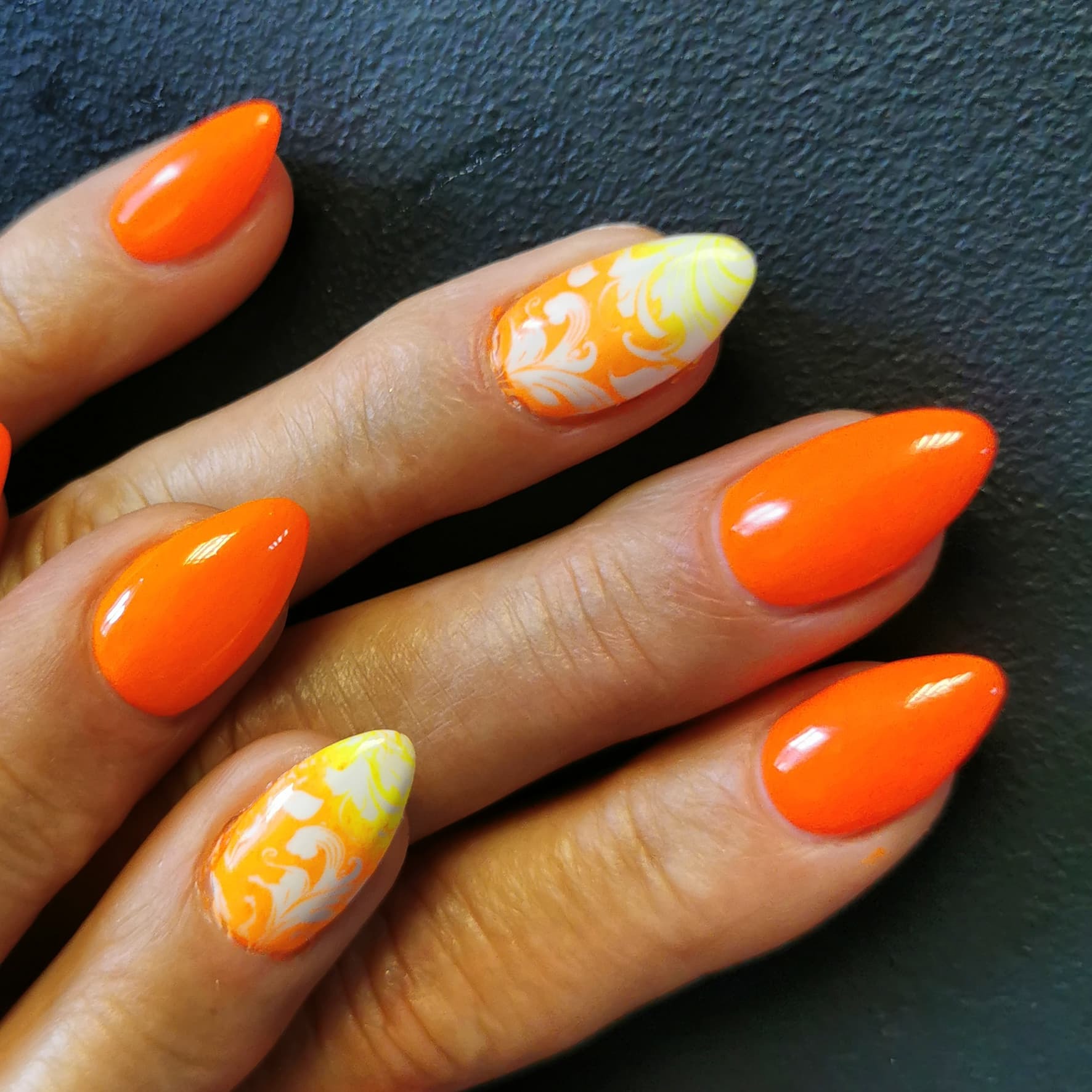 Dłoń z pomarańczowymi paznokciami o migdałowym kształcie, dwa z nich zdobione białym wzorem na pomarańczowo-żółtym tle.