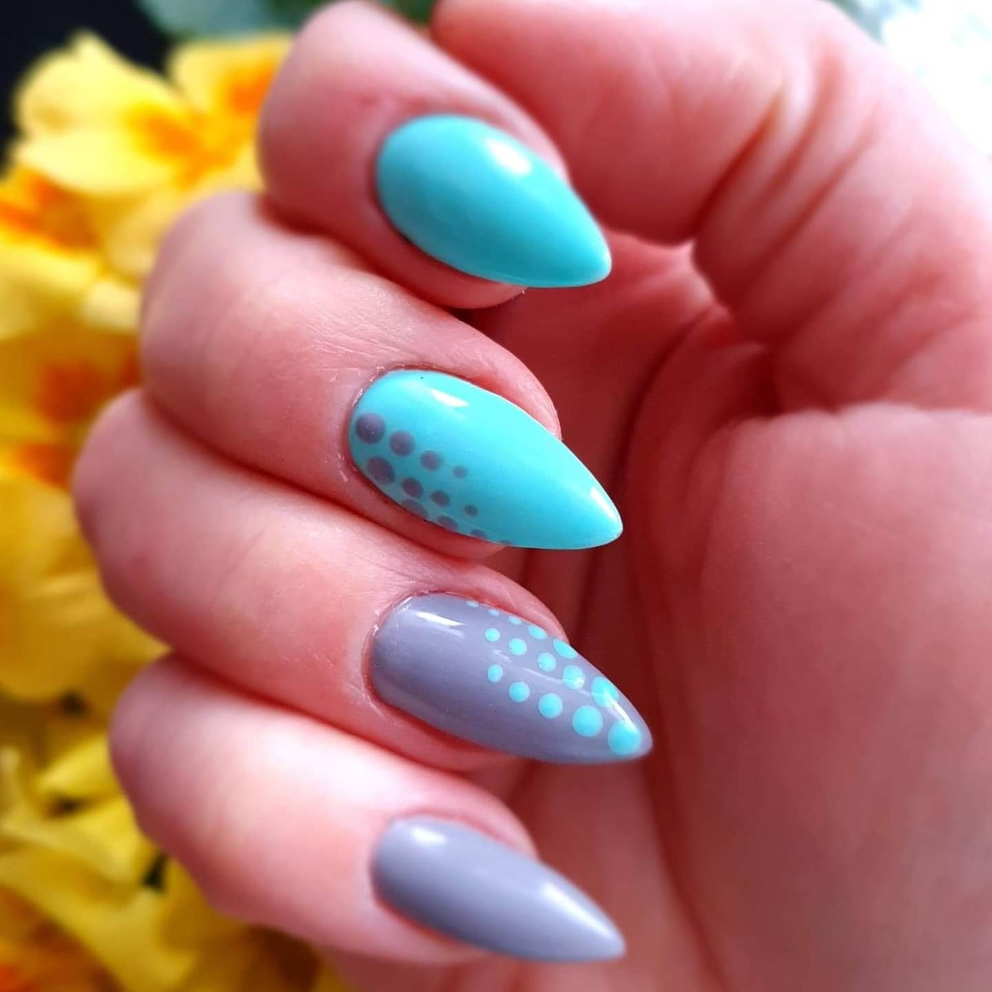 Dłoń z pomalowanymi paznokciami w kształcie migdałów, w kolorach turkusowym i szarym, zdobionymi kropkami; w tle rozmyte żółte kwiaty.