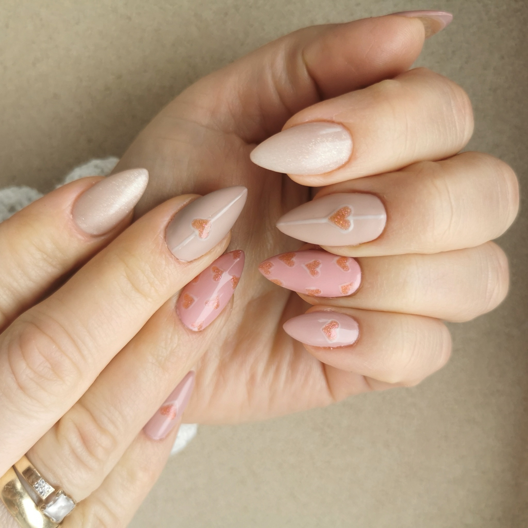 Dłonie z migdałowymi paznokciami w odcieniach różu i beżu, zdobione drobnymi, błyszczącymi serduszkami i geometrycznymi wzorami, złoty pierścionek z oczkiem na palcu.