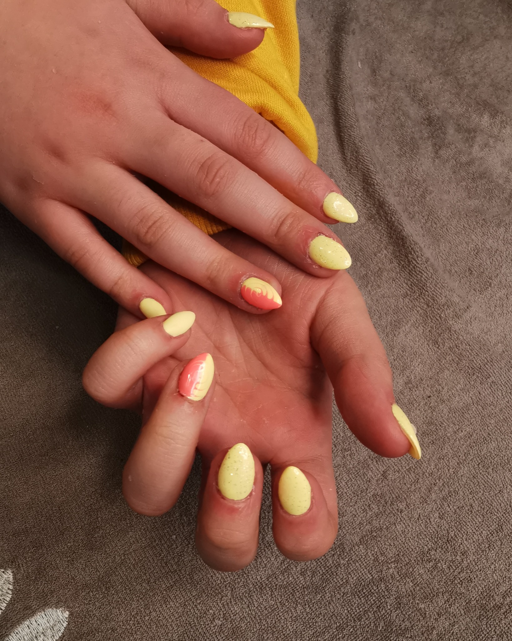 Dłonie z żółto-pomarańczowym manicure hybrydowym, część paznokci pomalowana w całości na żółto z brokatem, a część w połowie na żółto a w połowie na pomarańczowo z efektem ombre.