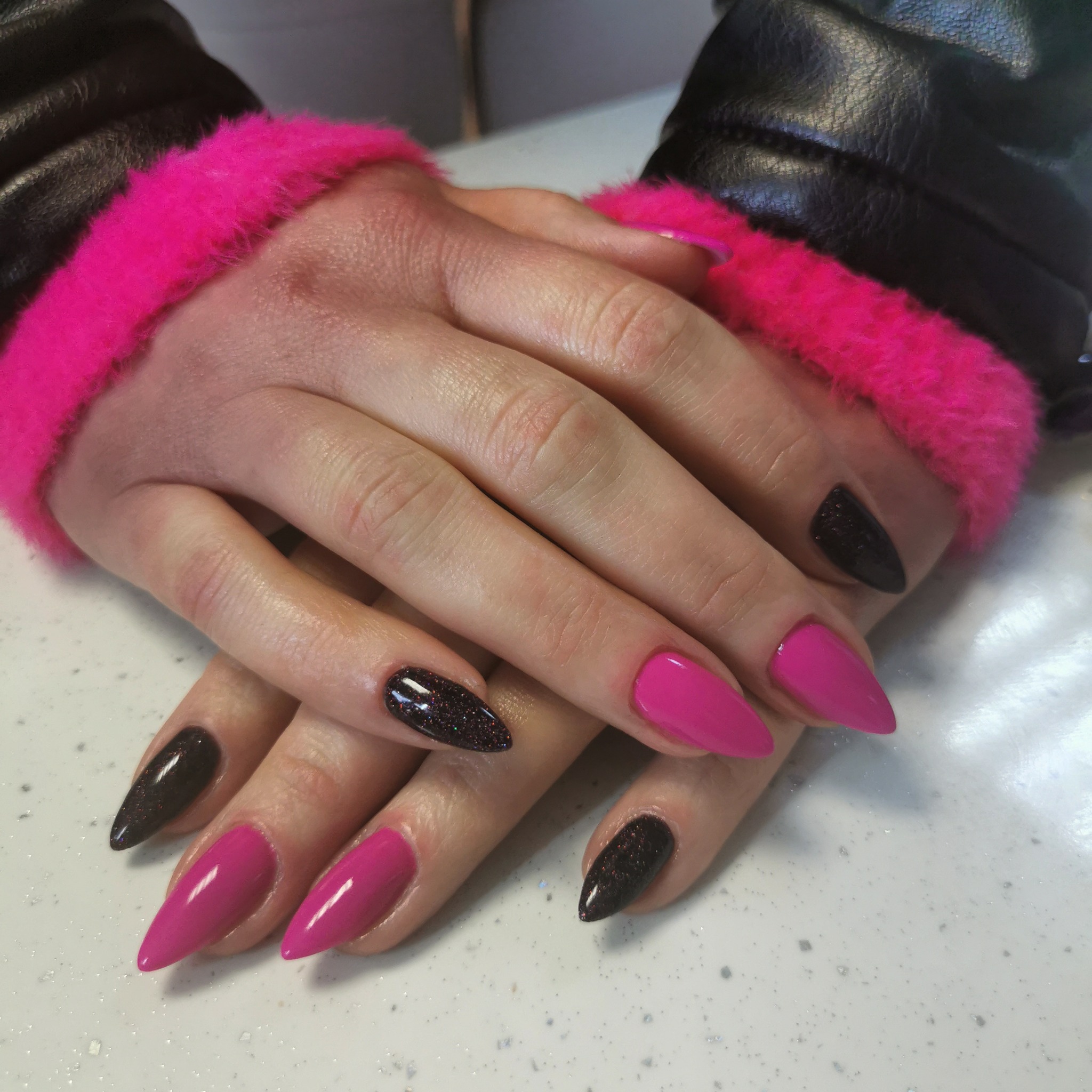 Dłonie z ostro zakończonymi paznokciami, pomalowane na różowo i ciemnobrązowo z brokatem, na jasnym blacie, w tle czarna kurtka z różowym futerkiem.