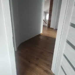 Widok na nowe panele podłogowe w korytarzu z białymi ścianami, widziane zza uchylonych drzwi z dekoracyjnymi wstawkami.