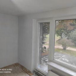 Dominik Miotk - Świeżo pomalowane na szaro wnętrze pokoju z widokiem na zewnątrz przez brudne okno, widoczne przygotowane podłoże pod parkiet lub panele.