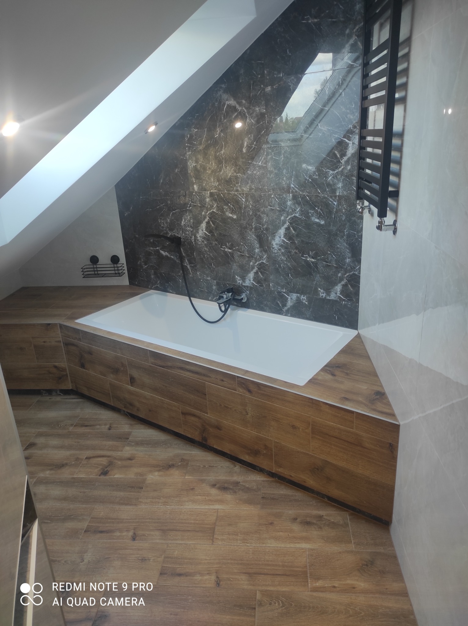 Nowoczesna łazienka na poddaszu z wanną obudowaną panelami drewnopodobnymi, marmurową ścianą i grzejnikiem drabinkowym.