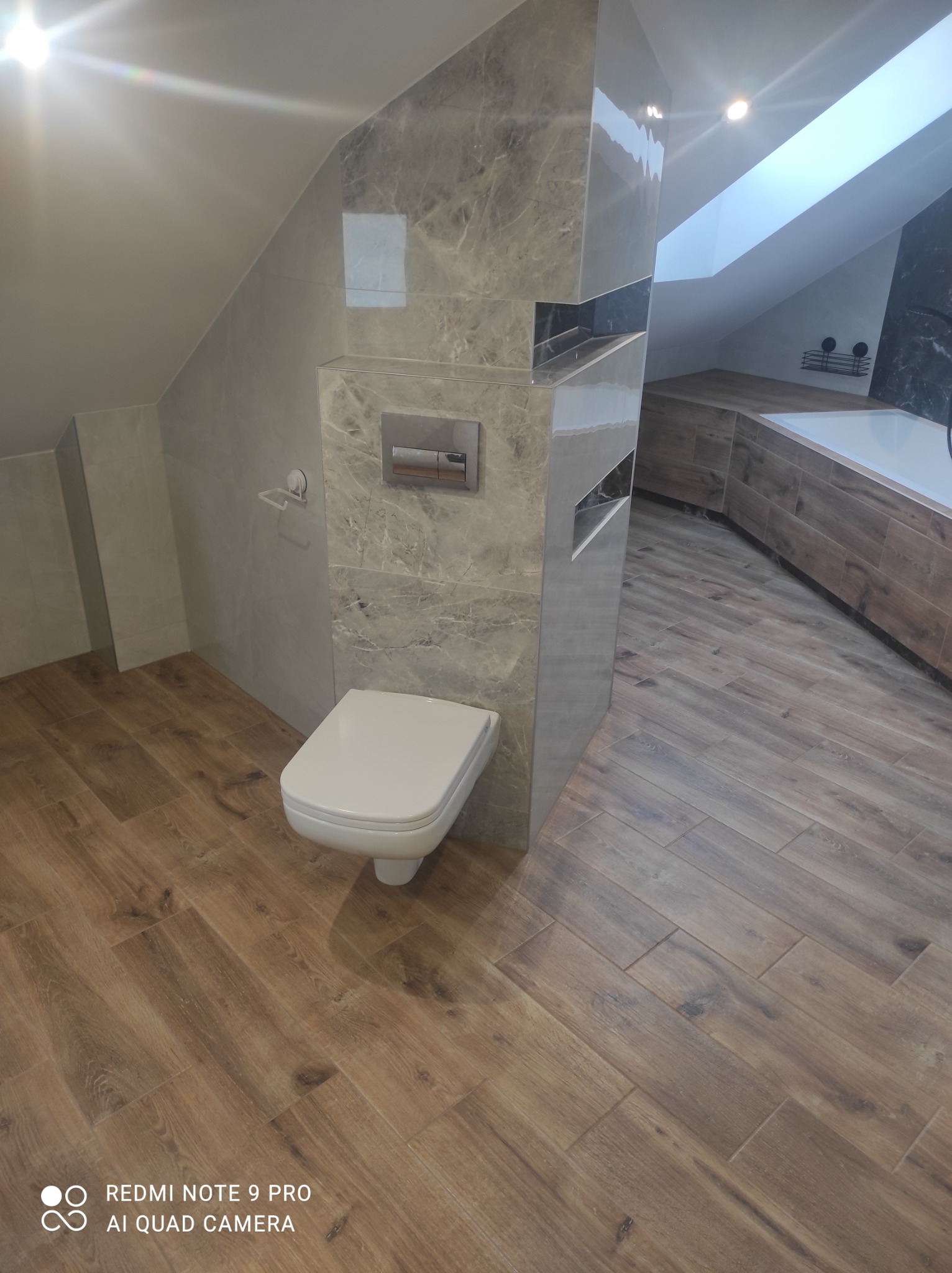 Nowoczesna łazienka na poddaszu z drewnopodobnymi płytkami na podłodze i ścianach w strefie wanny, marmurowe płytki na ścianach przy toalecie, biała wisząca toaleta i okno dachowe w suficie.