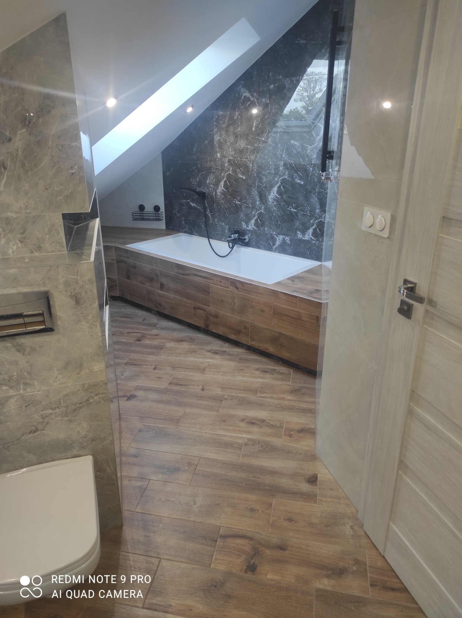 Nowoczesna łazienka na poddaszu z marmurowymi płytkami na ścianach, drewnopodobną podłogą i obudową wanny, oraz oknem dachowym w skosie.