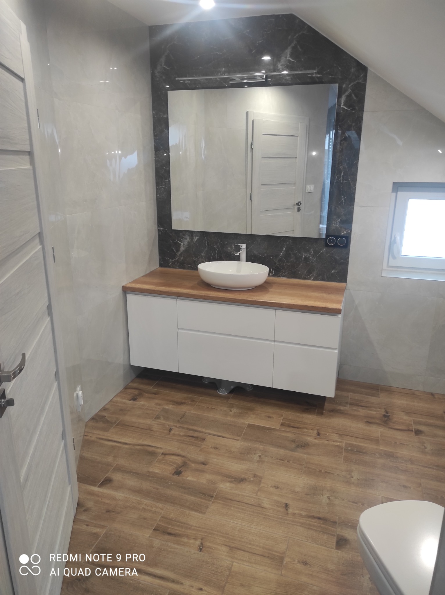 Nowoczesna łazienka na poddaszu z drewnopodobną podłogą, białą szafką pod umywalką z drewnianym blatem, okrągłą umywalką i lustrem w czarnej marmurowej ramie.