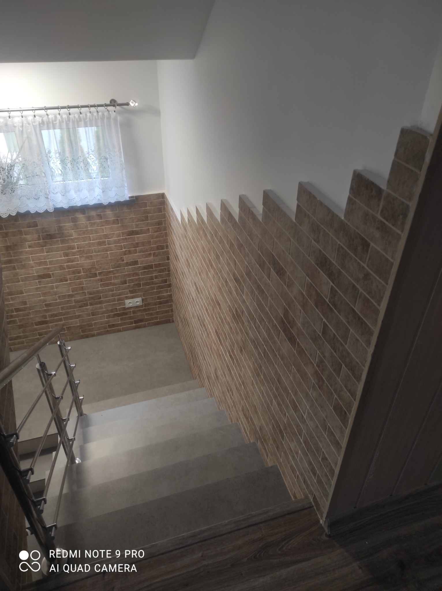 Klatka schodowa z nowymi płytkami imitującymi cegłę na ścianie do połowy wysokości, szare schody i drewnopodobne panele na podłodze.