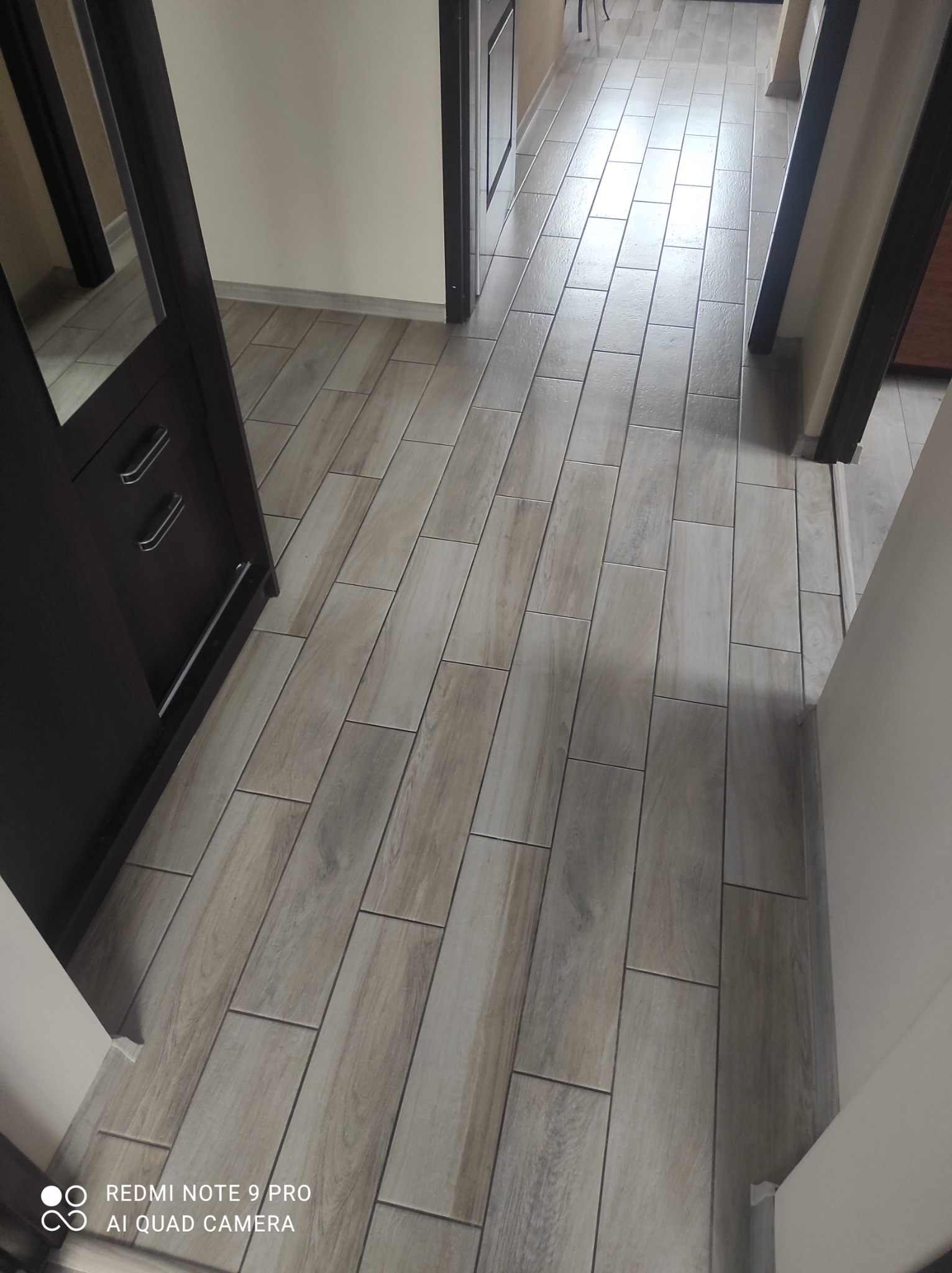 Korytarz wyłożony podłużnymi płytkami imitującymi drewno, widok z góry, ciemna szafka z lustrem po lewej stronie.