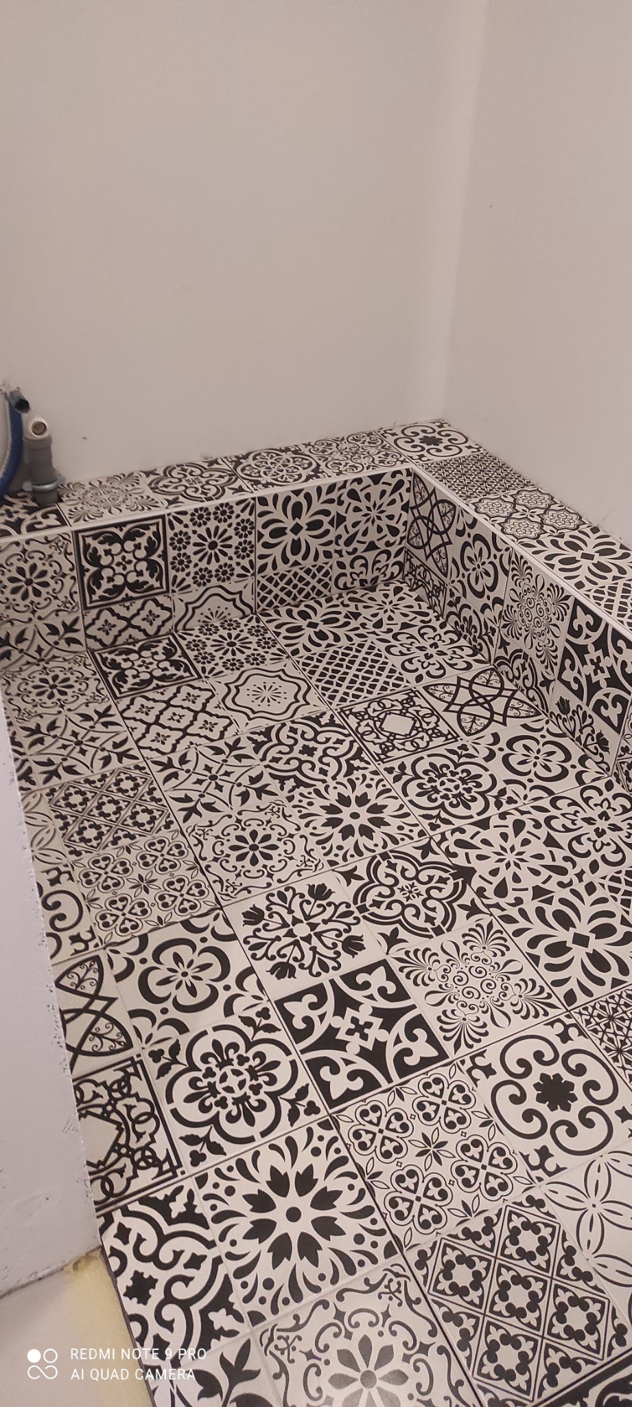 Czarno-białe, wzorzyste płytki ceramiczne ułożone na podłodze i ściankach brodzika, widoczne przyłącze wodne.