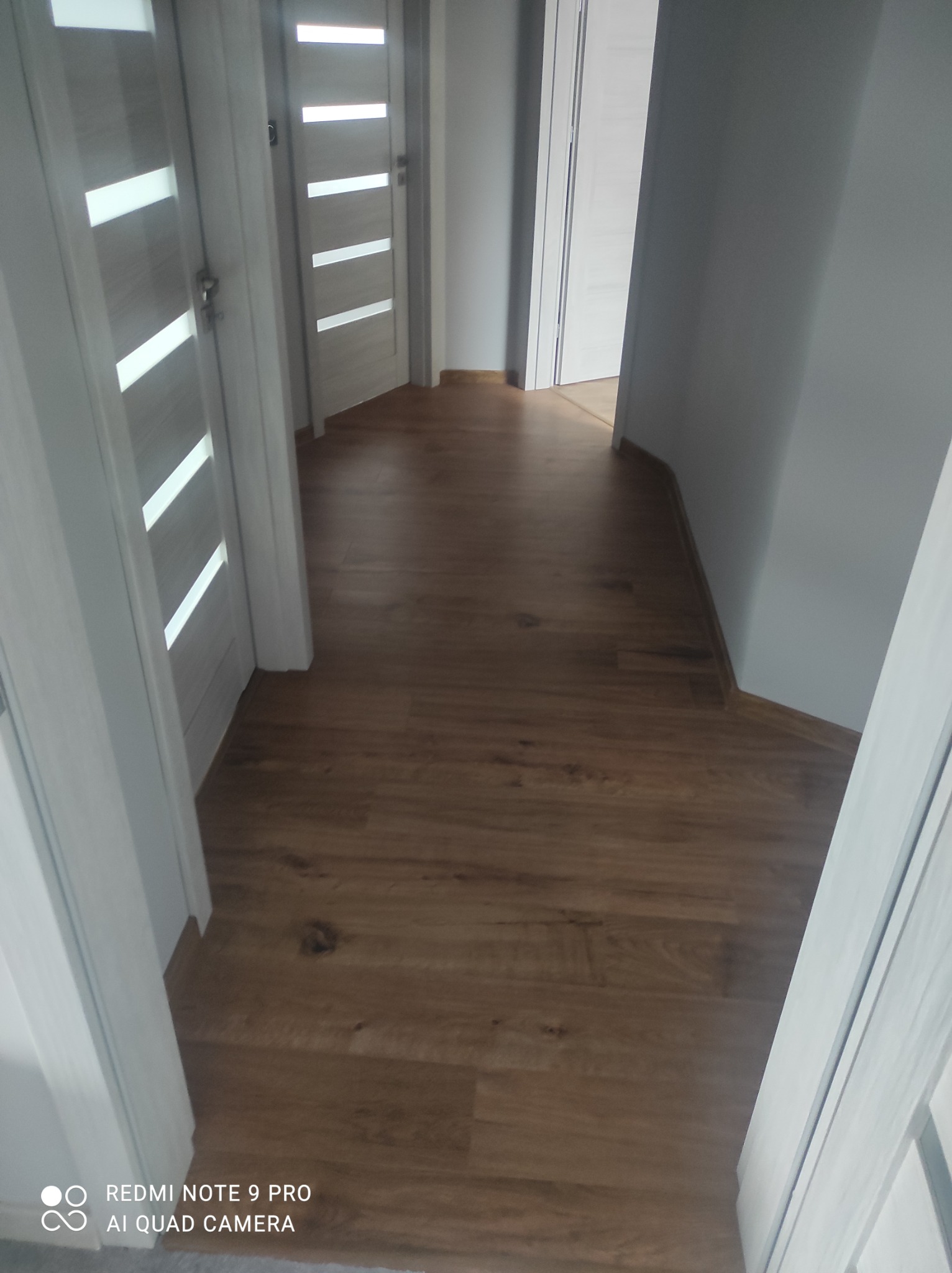 Korytarz z nową, brązową podłogą imitującą drewno, widoczne białe drzwi z poziomymi przeszkleniami i szare ściany.