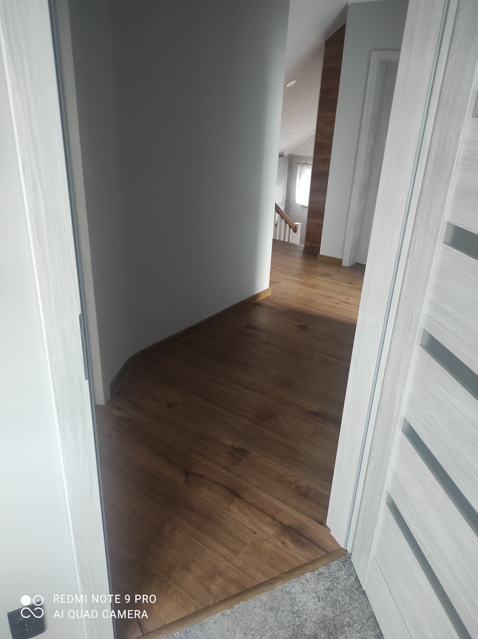 Widok na nowe panele podłogowe w korytarzu z białymi ścianami, widziane zza uchylonych drzwi z dekoracyjnymi wstawkami.