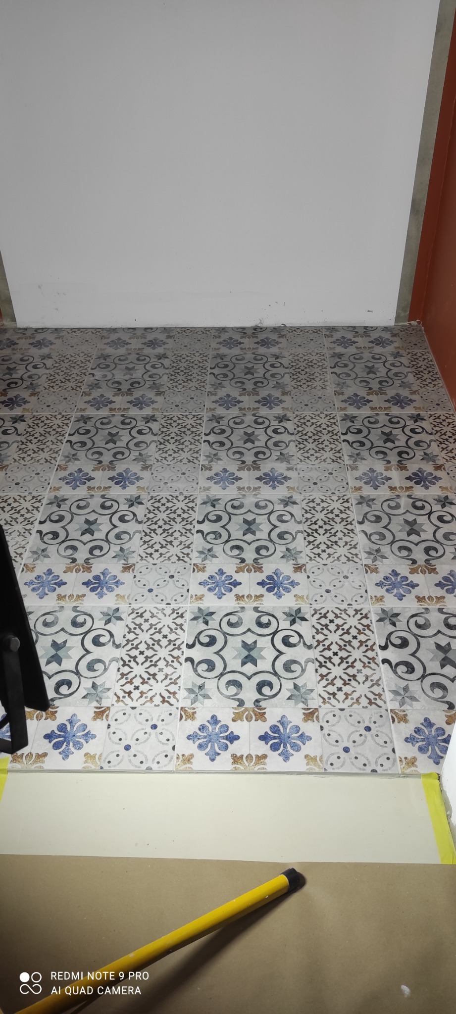 Ułożona podłoga z płytek o wzorze patchworkowym w odcieniach szarości, błękitu i brązu, widoczna z góry, z fragmentem żółtej taśmy malarskiej i narzędzia przy krawędzi