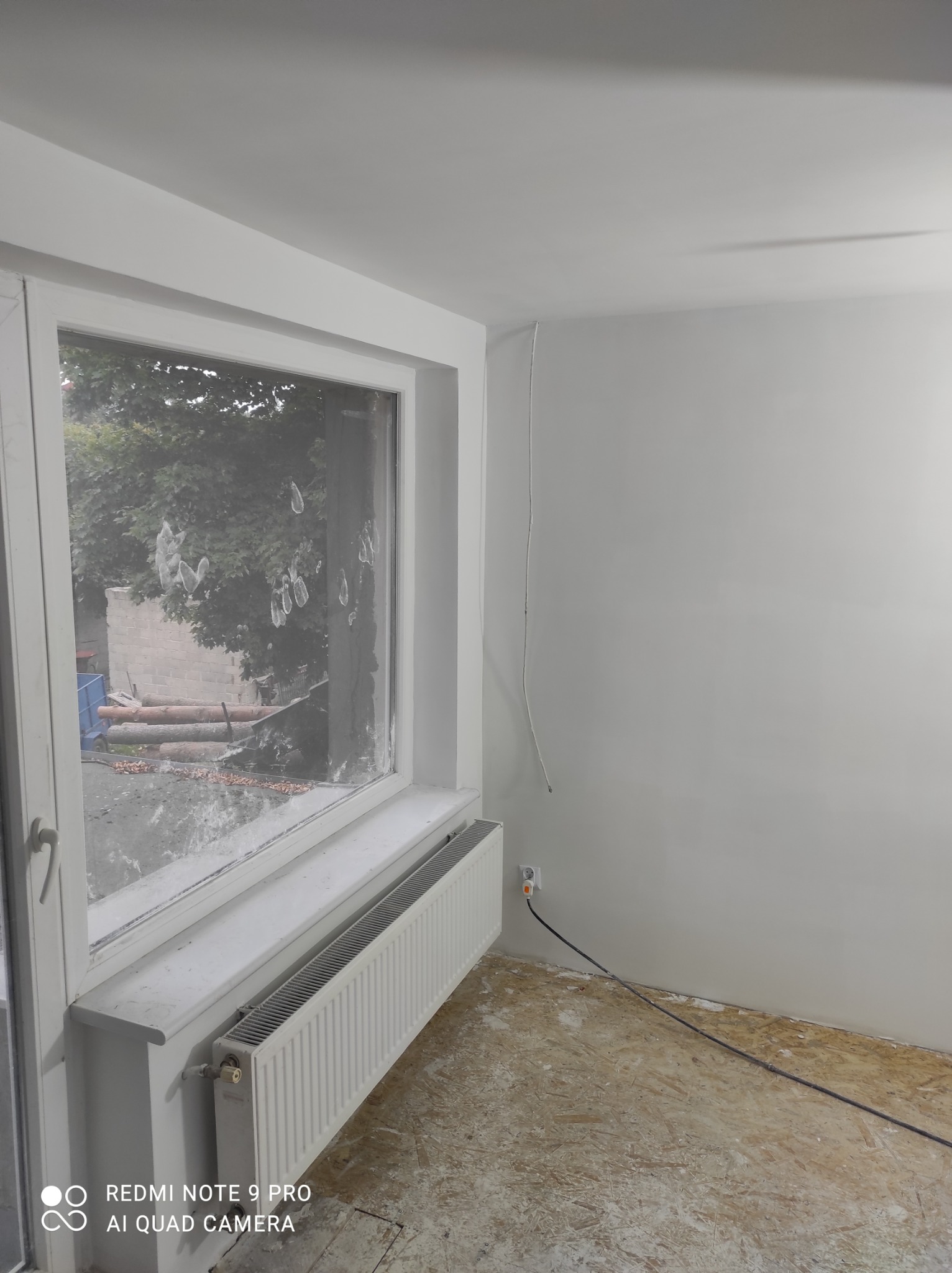 Wnętrze pokoju po malowaniu ścian na biało, widoczne białe ramy okienne z zabrudzeniami po farbie, grzejnik pod oknem, surowa podłoga z płyty OSB i wystający kabel elektryczny ze ściany.