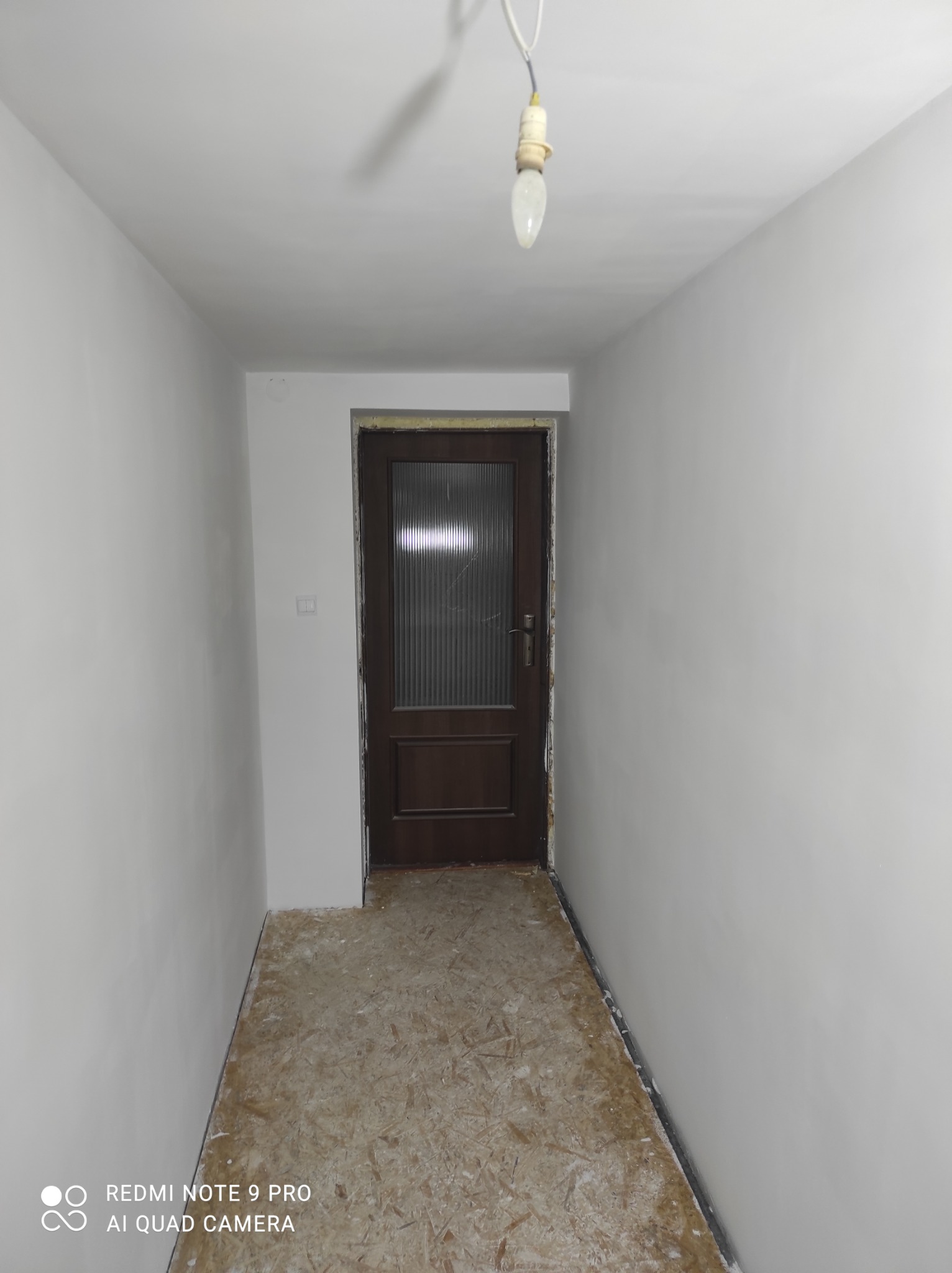 Korytarz w trakcie remontu: świeżo pomalowane ściany na biało, surowa podłoga z płyt OSB, ciemnobrązowe drzwi z szybą i wisząca żarówka.