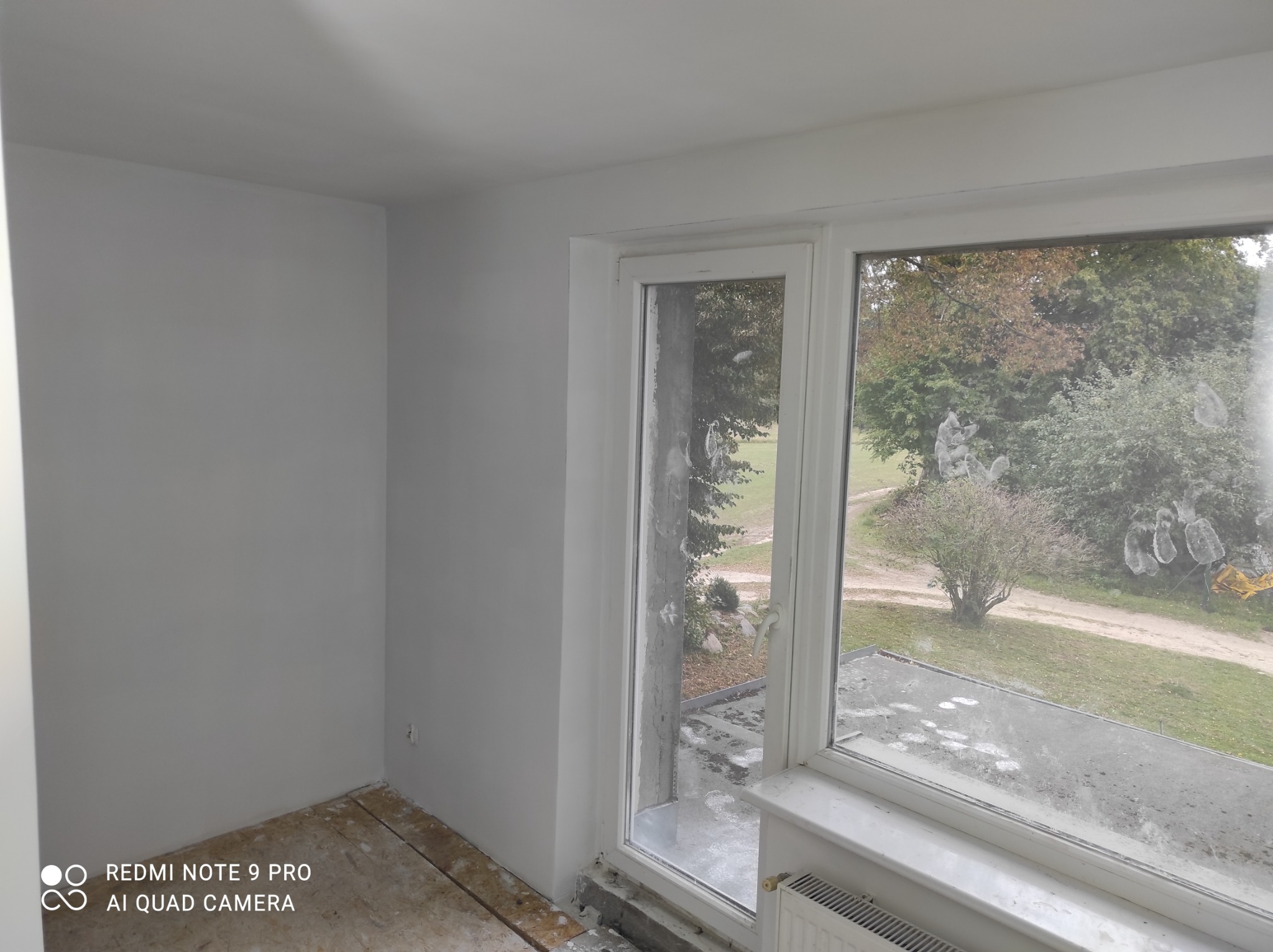 Świeżo pomalowane na szaro wnętrze pokoju z widokiem na zewnątrz przez brudne okno, widoczne przygotowane podłoże pod parkiet lub panele.