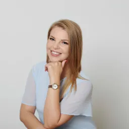 Uśmiechnięta blondynka w błękitnej bluzce i złotym zegarku, oparta podbródkiem o dłoń na jasnym tle.