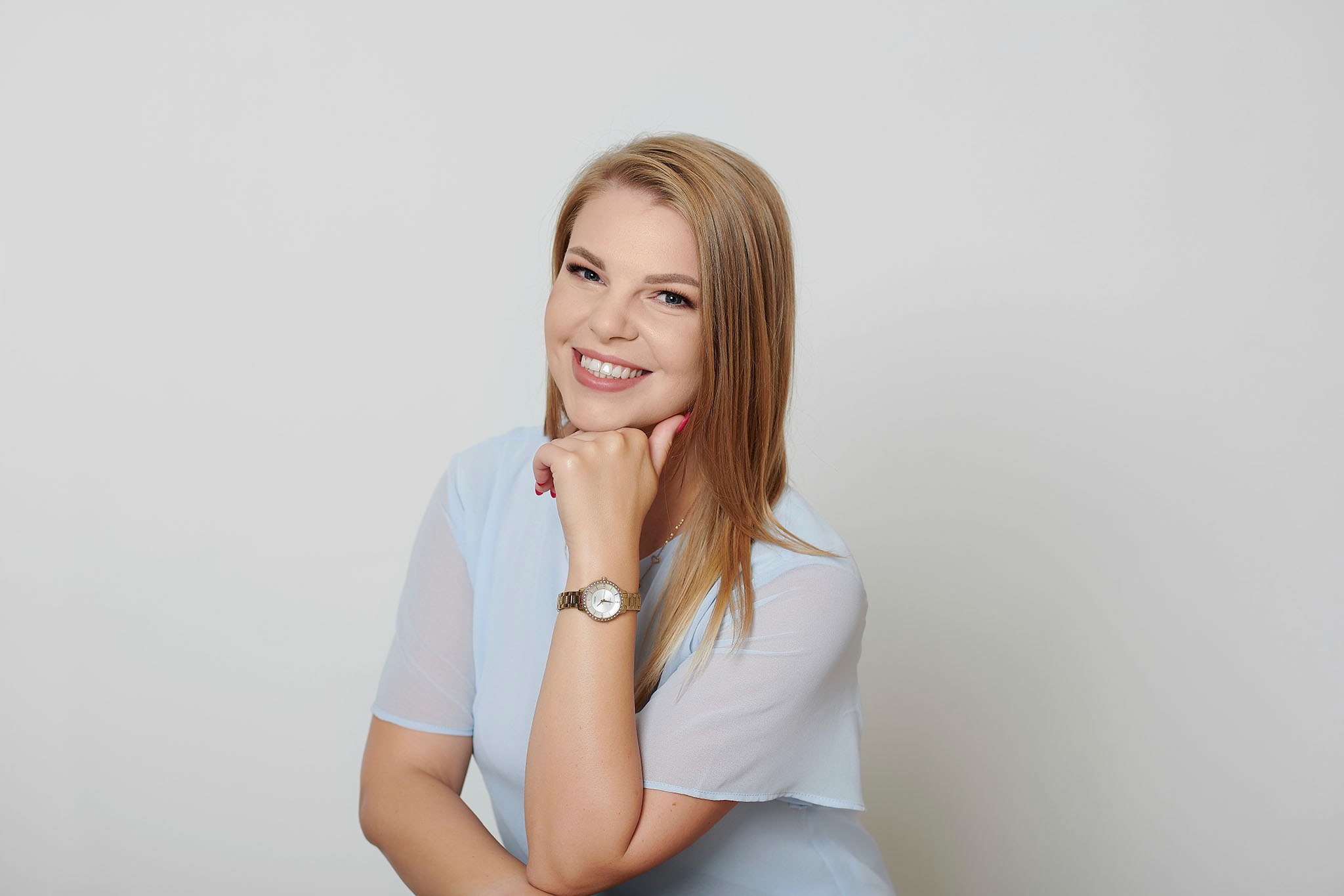Uśmiechnięta blondynka w błękitnej bluzce i złotym zegarku, oparta podbródkiem o dłoń na jasnym tle.
