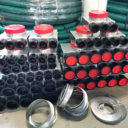 Magazynowane elementy systemu rekuperacji: skrzynki rozdzielcze z czarnymi króćcami i czerwonymi pokrywami, zwój zielonych rur wentylacyjnych oraz metalowe obejmy mocujące.