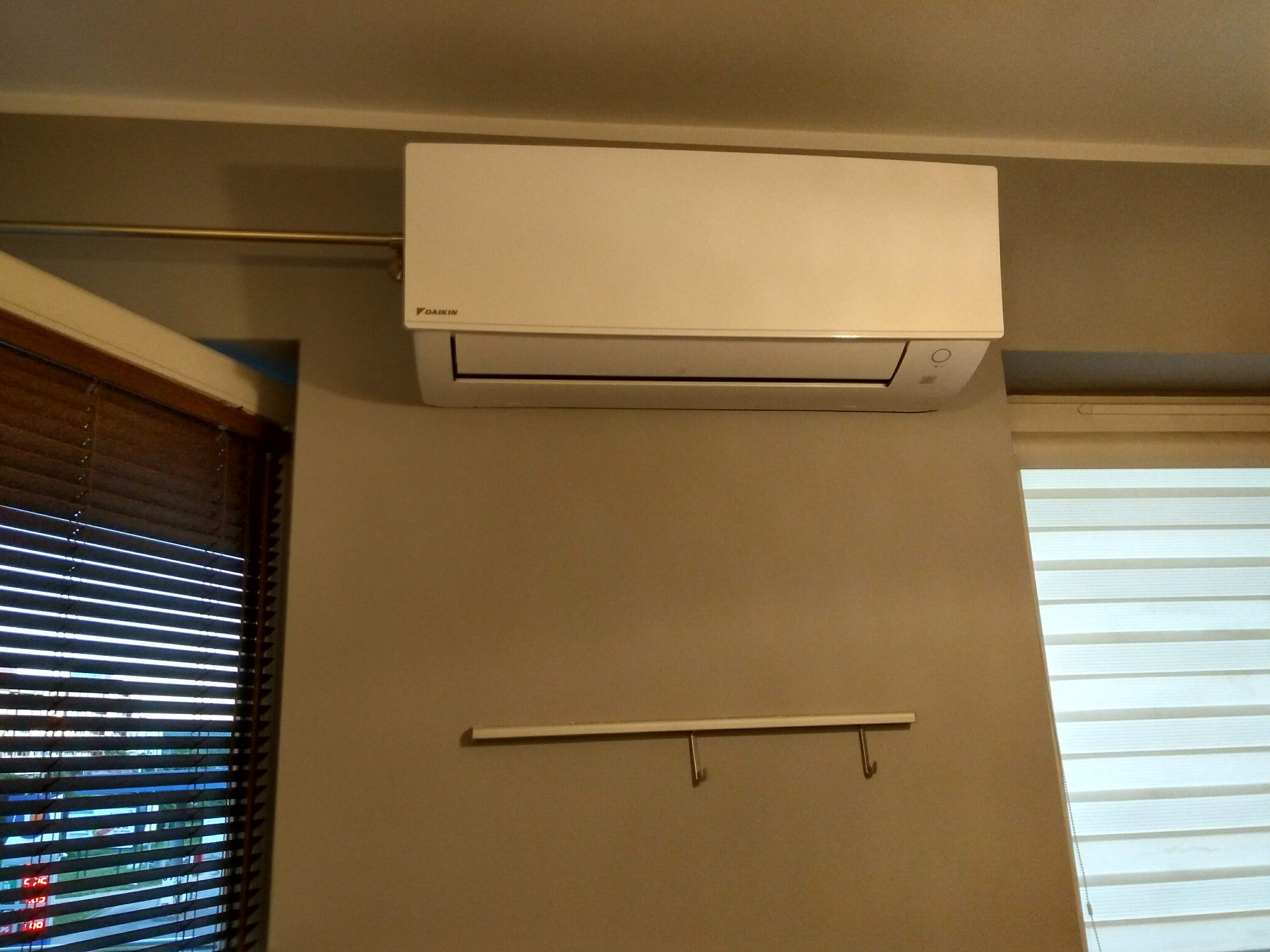 Biały klimatyzator Daikin zamontowany na ścianie w pomieszczeniu z roletami poziomymi, widoczne elementy instalacji i mały wieszak.