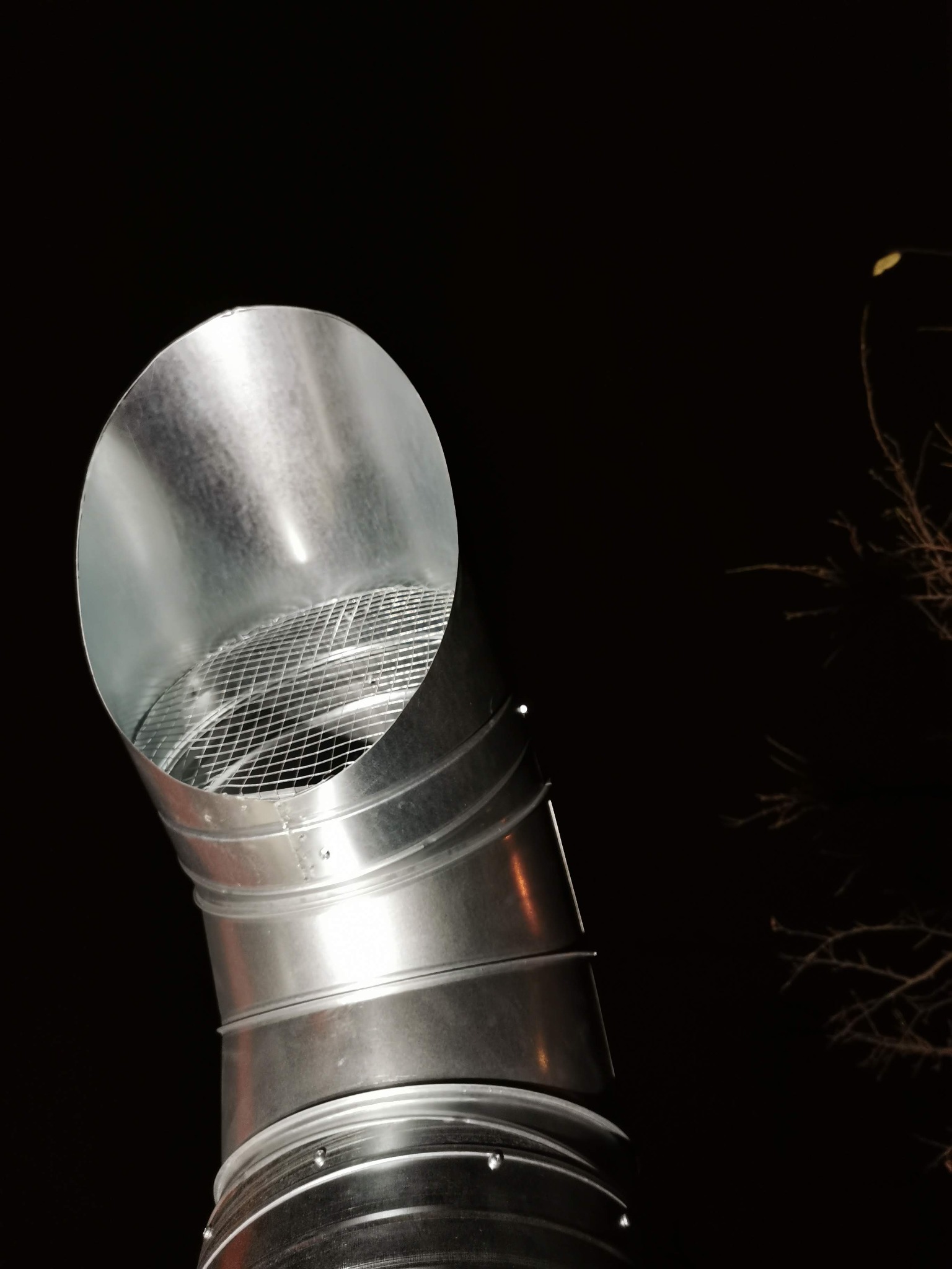 Srebrny, metalowy przewód wentylacyjny z siatką zabezpieczającą, skierowany w górę na tle nocnego nieba i gałęzi drzew