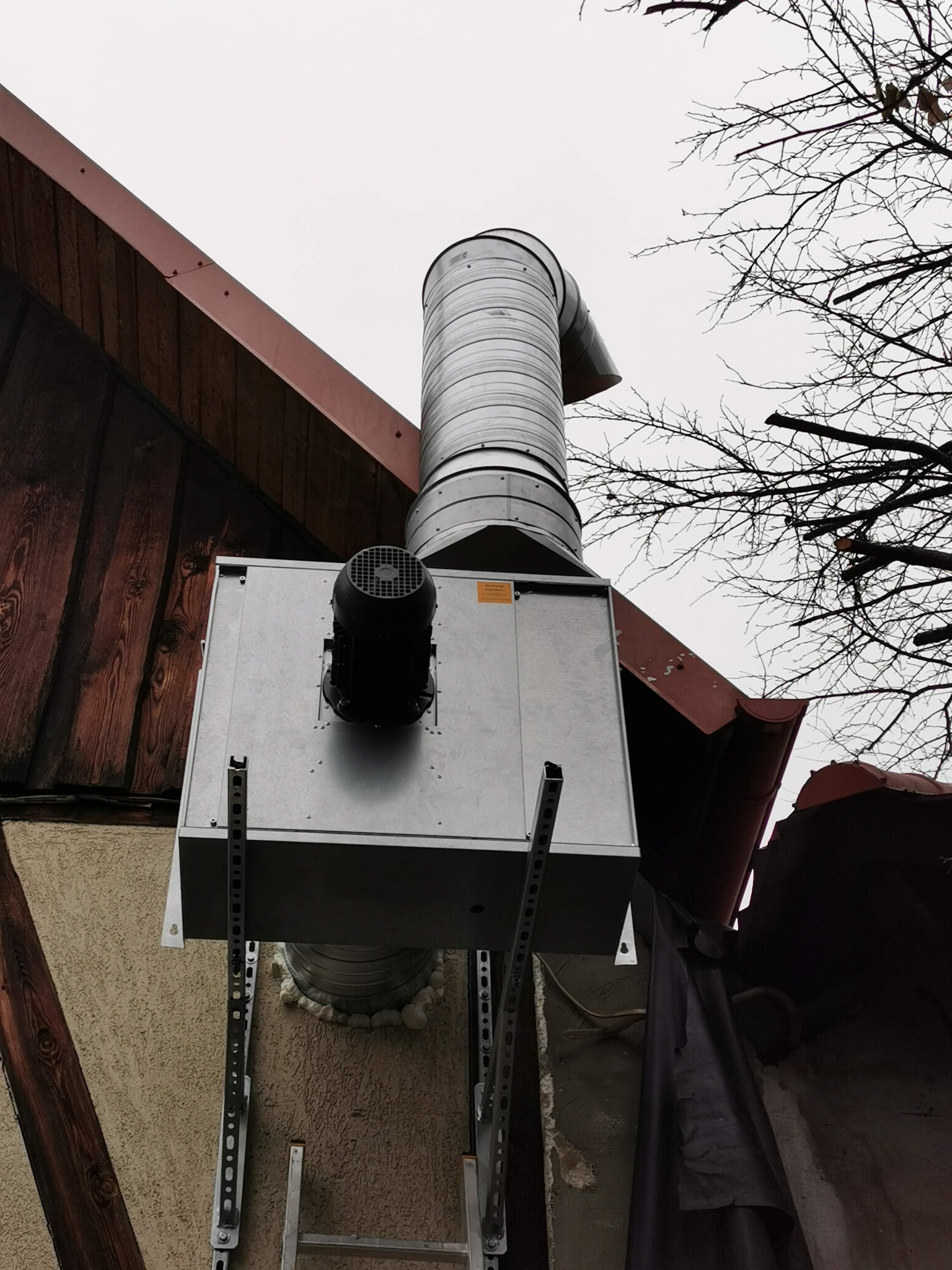 Zewnętrzna instalacja rekuperacji z wentylatorem na ścianie budynku z drewnianą konstrukcją dachu i gałęziami drzew na tle pochmurnego nieba.