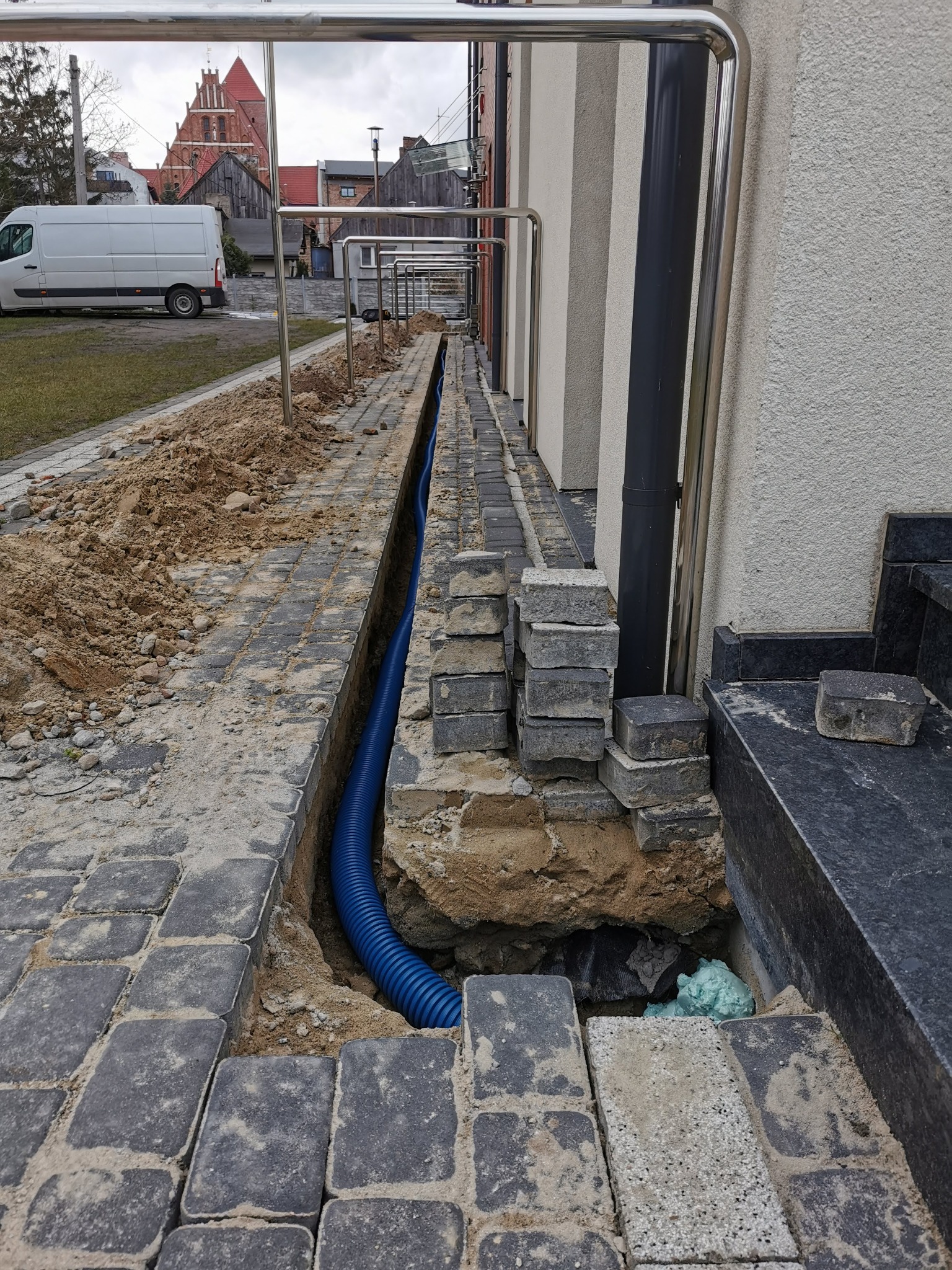 Wkopany niebieski karbowany przewód w wąskim wykopie wzdłuż chodnika z kostki, przygotowanie do instalacji, w tle budynek z czerwonej cegły i biały samochód dostawczy.