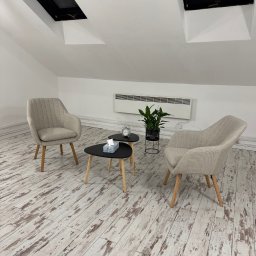 Przyjazna Przestrzeń Agata Pizoń - Jasne, minimalistyczne wnętrze gabinetu z dwoma fotelami, stolikiem z chusteczkami i zegarem, oraz rośliną w czarnej doniczce, doświetlone przez okna dachowe.