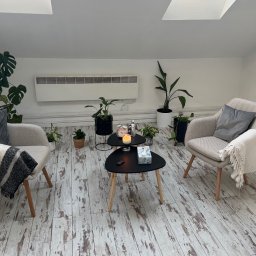 Przyjazna Przestrzeń Agata Pizoń - Jasne, minimalistyczne wnętrze gabinetu z dwoma fotelami, stolikami kawowymi, roślinami doniczkowymi i drewnianą podłogą. Na stoliku świeca i pudełko chusteczek.