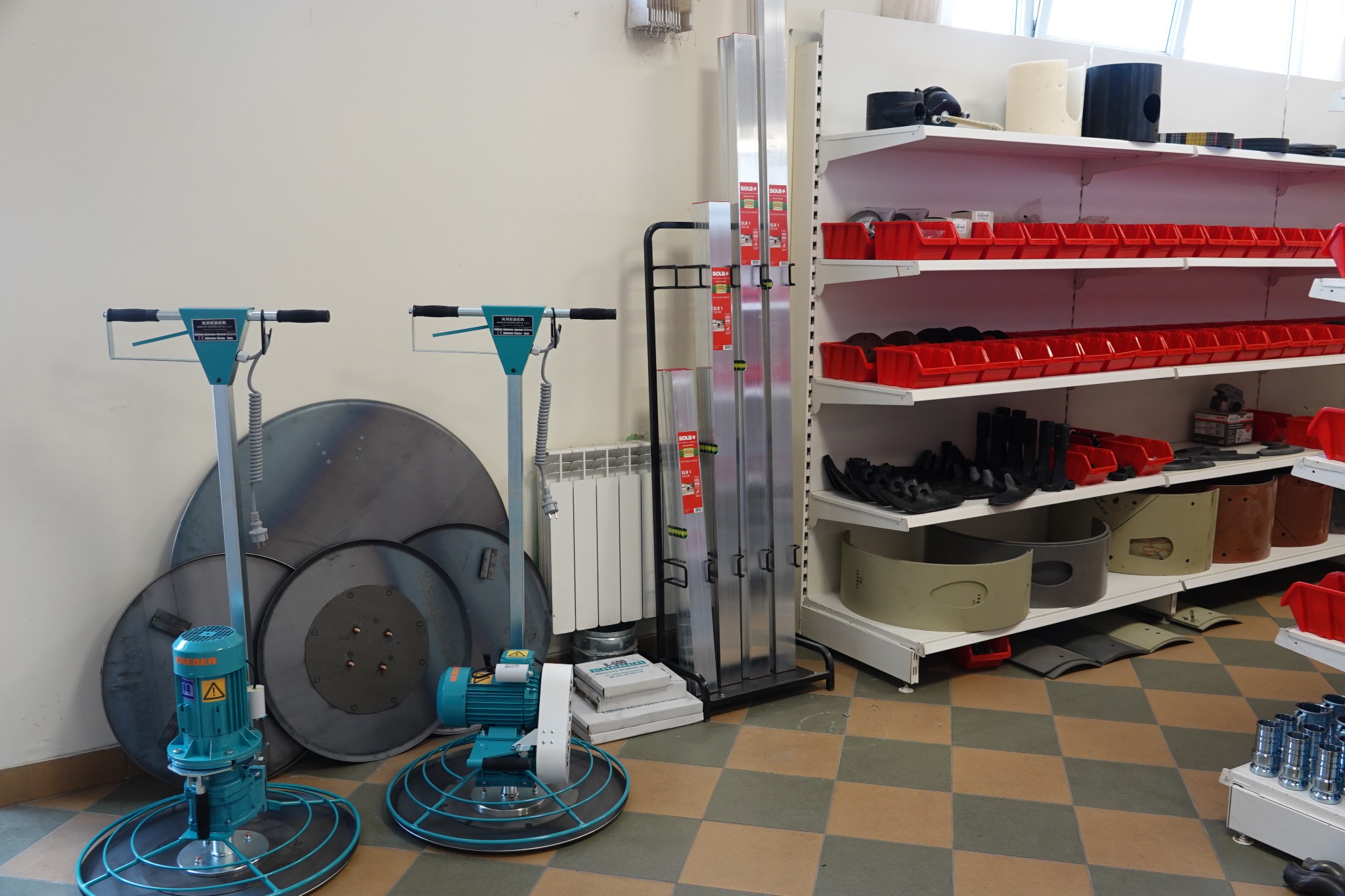 Dwie zacieraczki do betonu marki Kreber ustawione w pomieszczeniu obok regałów z narzędziami i akcesoriami budowlanymi.