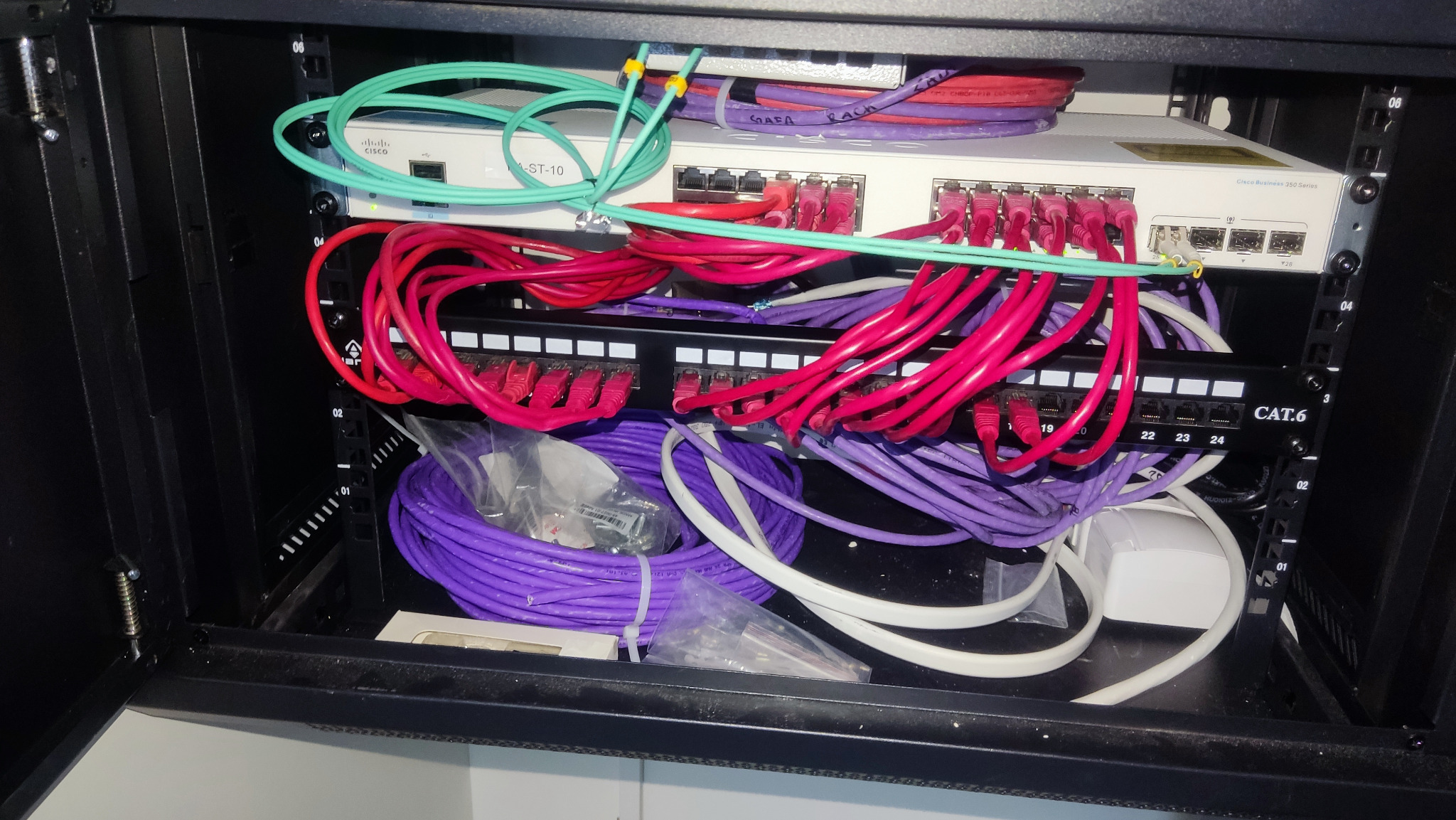 Szafa rack z zainstalowanym switchem Cisco, panelem krosowym CAT.6 i okablowaniem w kolorach: czerwonym, fioletowym, białym i zielonym. Widoczne oznaczenia portów na panelu krosowym.