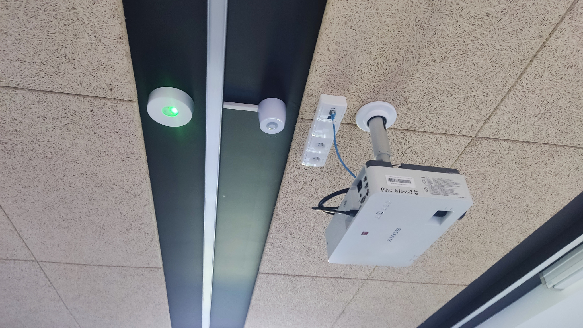 Sufit podwieszany z zamontowanym projektorem Sony, czujnikiem ruchu z zieloną diodą, gniazdem elektrycznym i listwą oświetleniową LED.