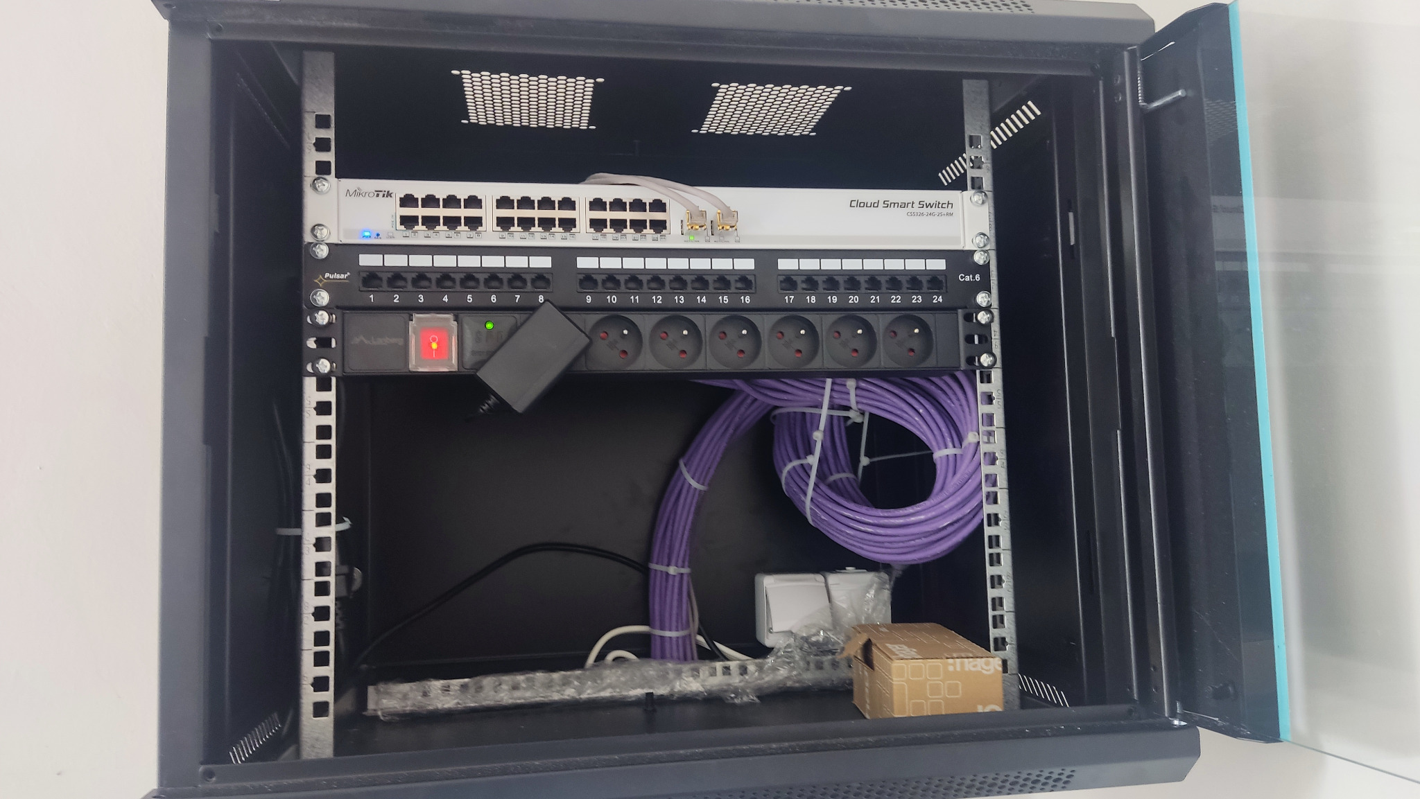Szafa rack z zamontowanym switchem Mikrotik Cloud Smart Switch, panelem krosowniczym Cat.6, listwą zasilającą Pulsar oraz uporządkowanymi kablami sieciowymi w kolorze fioletowym.