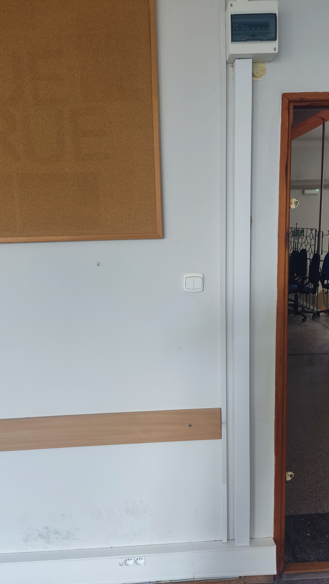 Biała ściana z tablicą korkową, włącznikiem światła, listwą przypodłogową z gniazdkami, drewnianą listwą na ścianie i skrzynką elektryczną pod sufitem, widok na salę przez otwarte drzwi.