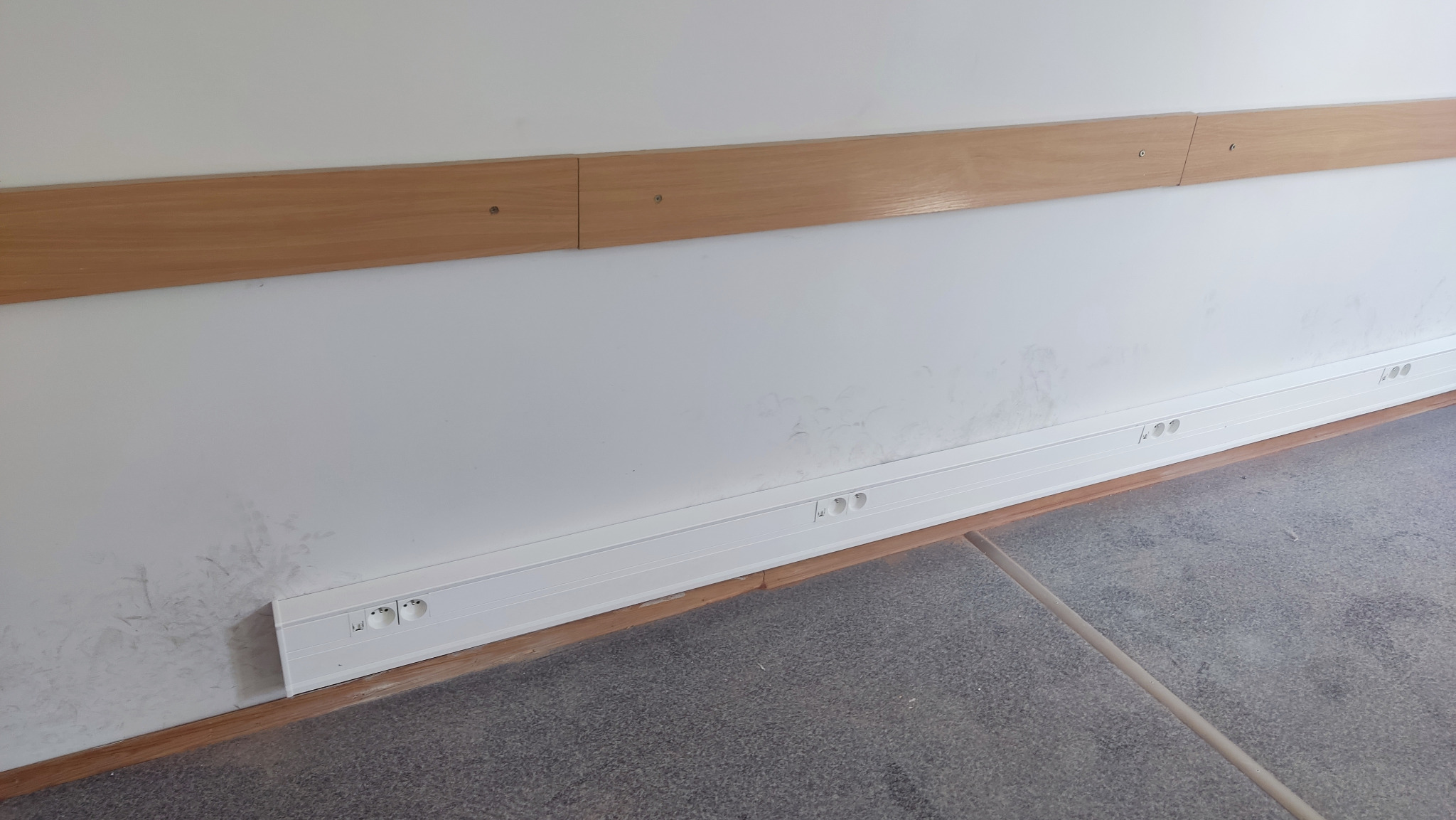 Biała listwa przypodłogowa z gniazdkami elektrycznymi zamontowana wzdłuż ściany z drewnianą poręczą, widoczne ślady zabrudzeń na ścianie.