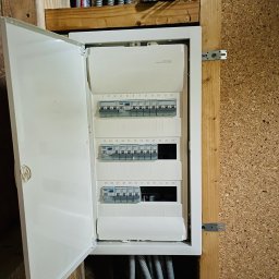 Wszystko w czasie - Otwarta, biała skrzynka elektryczna z bezpiecznikami, zamontowana na drewnianej ścianie. Widoczne kable w peszlach. U góry puszka Coca-Coli dla skali.