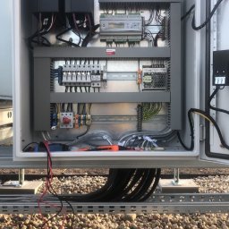 Wszystko w czasie - Otwarta szafa sterownicza z widocznymi elementami instalacji elektrycznej, takimi jak sterowniki VLT, bezpieczniki i okablowanie. Widoczne narzędzia w dolnej części szafy.